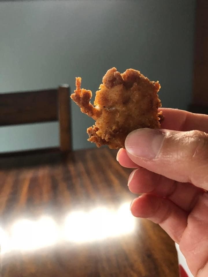 A hand holding a chicken nugget shaped like a cartoon character, with a wooden table and chair in the background