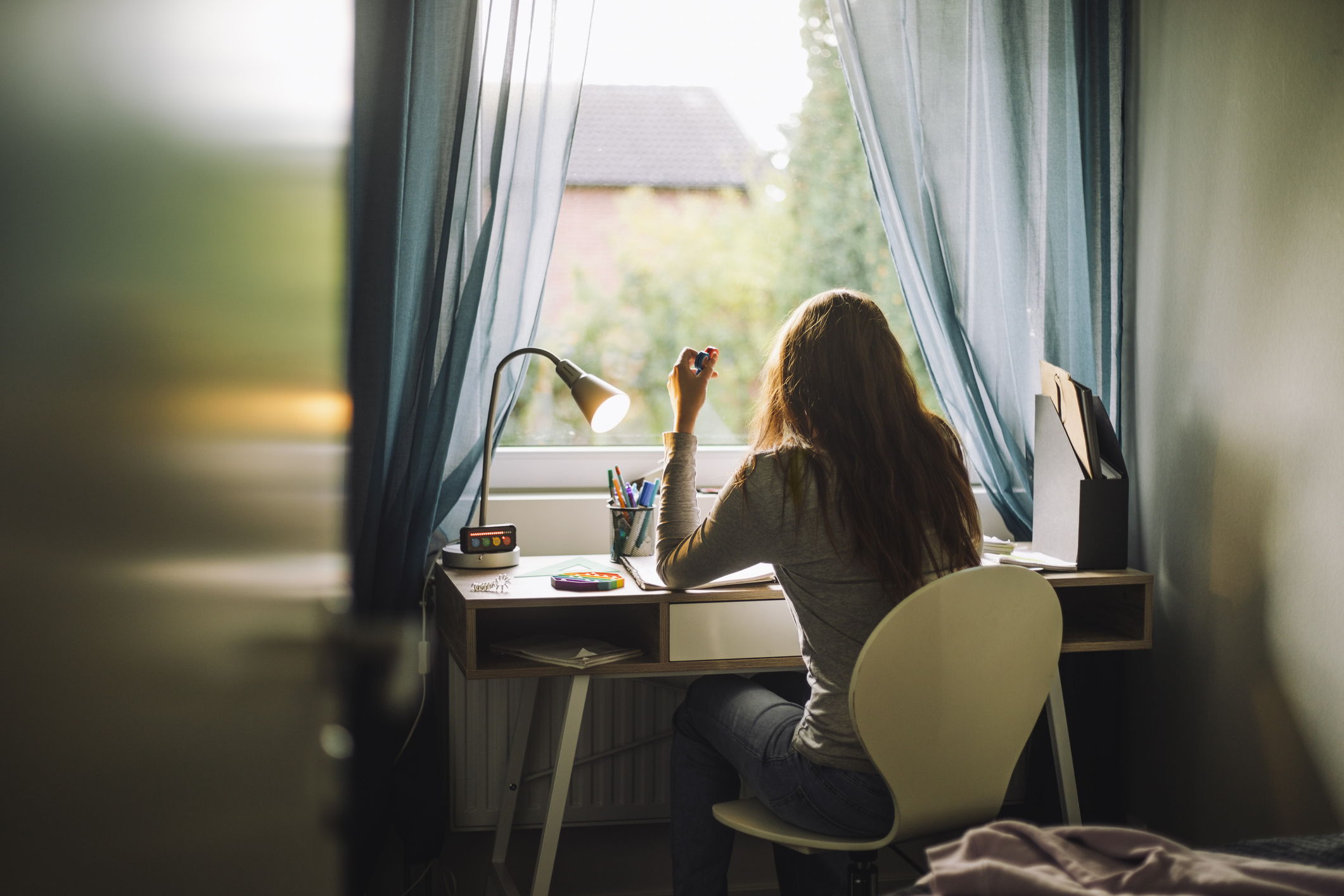 A person with long hair sits at a desk by a window, holding something up while working. Sunlight filters through the curtains into the room