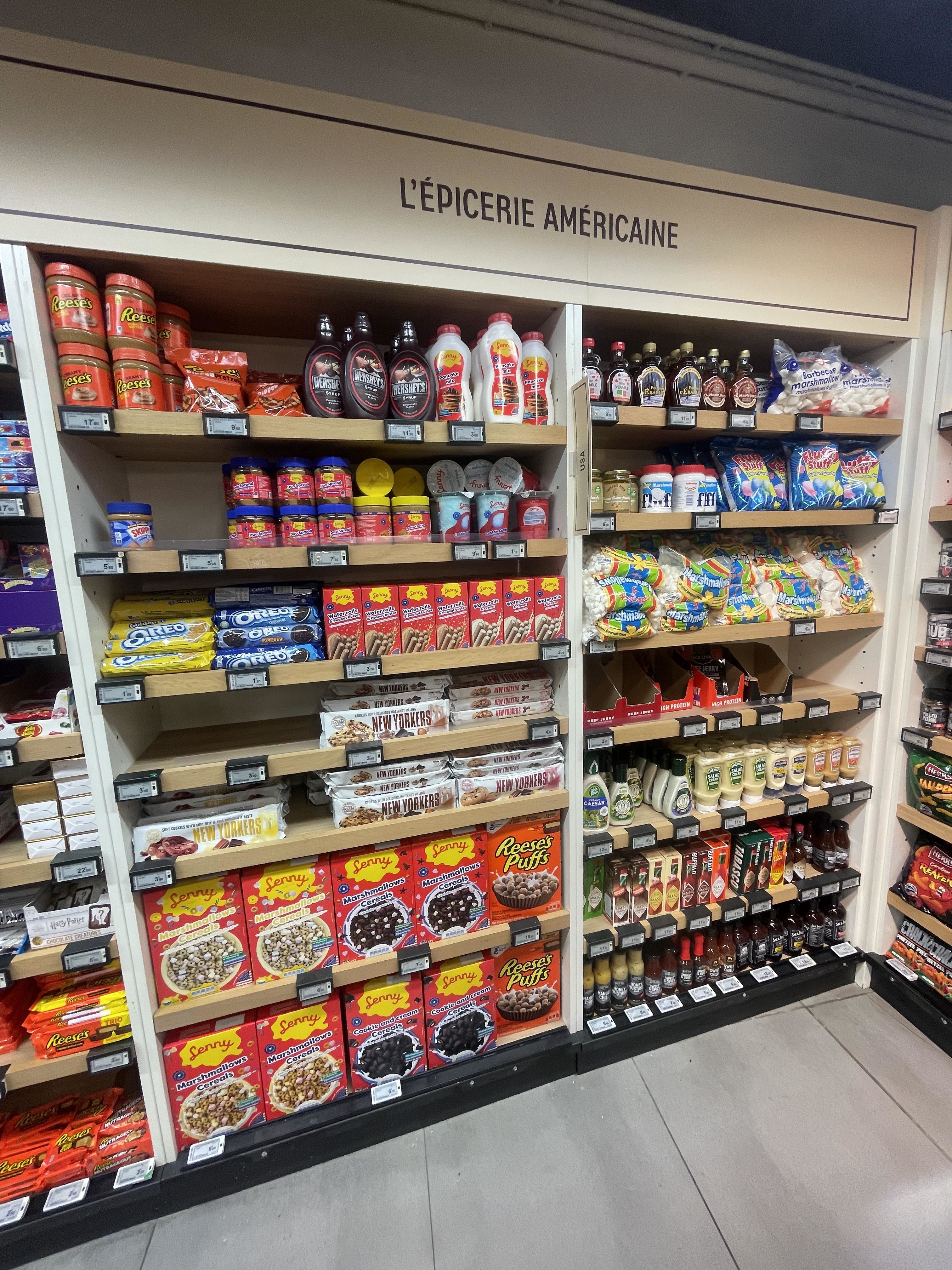 Grocery store aisle labeled "L'ÉPICERIE AMÉRICAINE" with various American food products like cereals, syrup, candy, and sauces on display