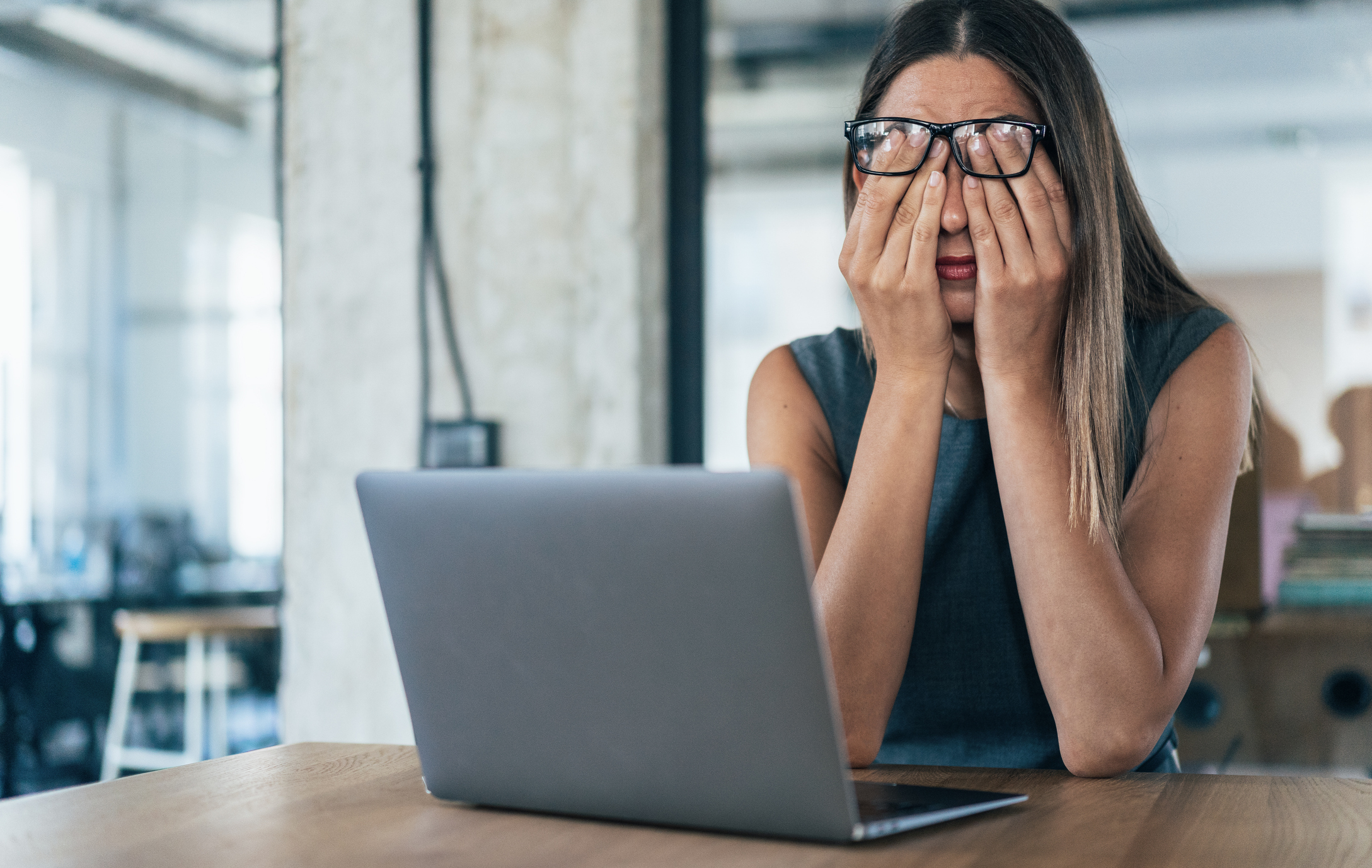 A person in casual clothing sits at a desk with a laptop, rubbing their eyes in apparent frustration or fatigue