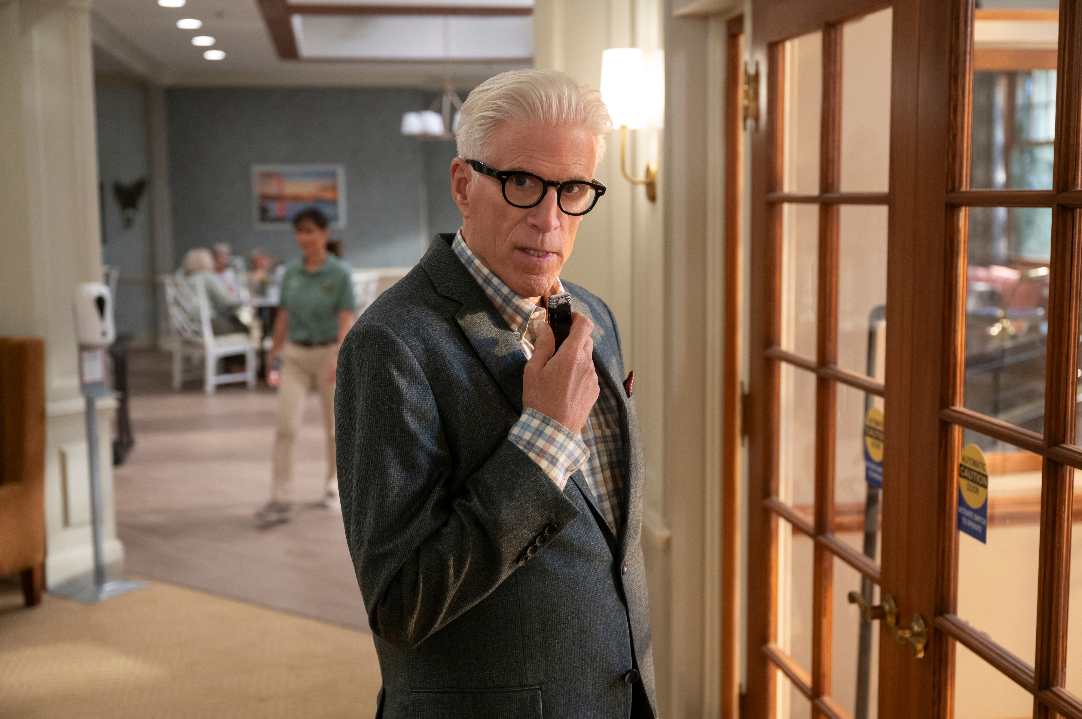 Ted Danson wearing a suit with glasses, posing indoors, standing near a wooden glass door, with people blurred in the background