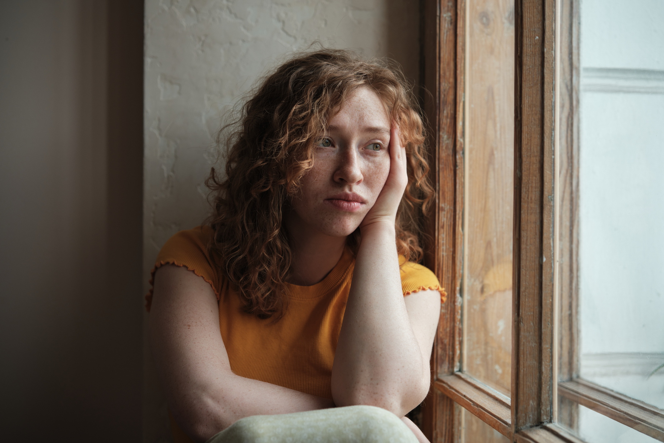 A woman with curly hair wearing a short-sleeved top leans on a windowsill, looking out thoughtfully, with her head resting on her hand