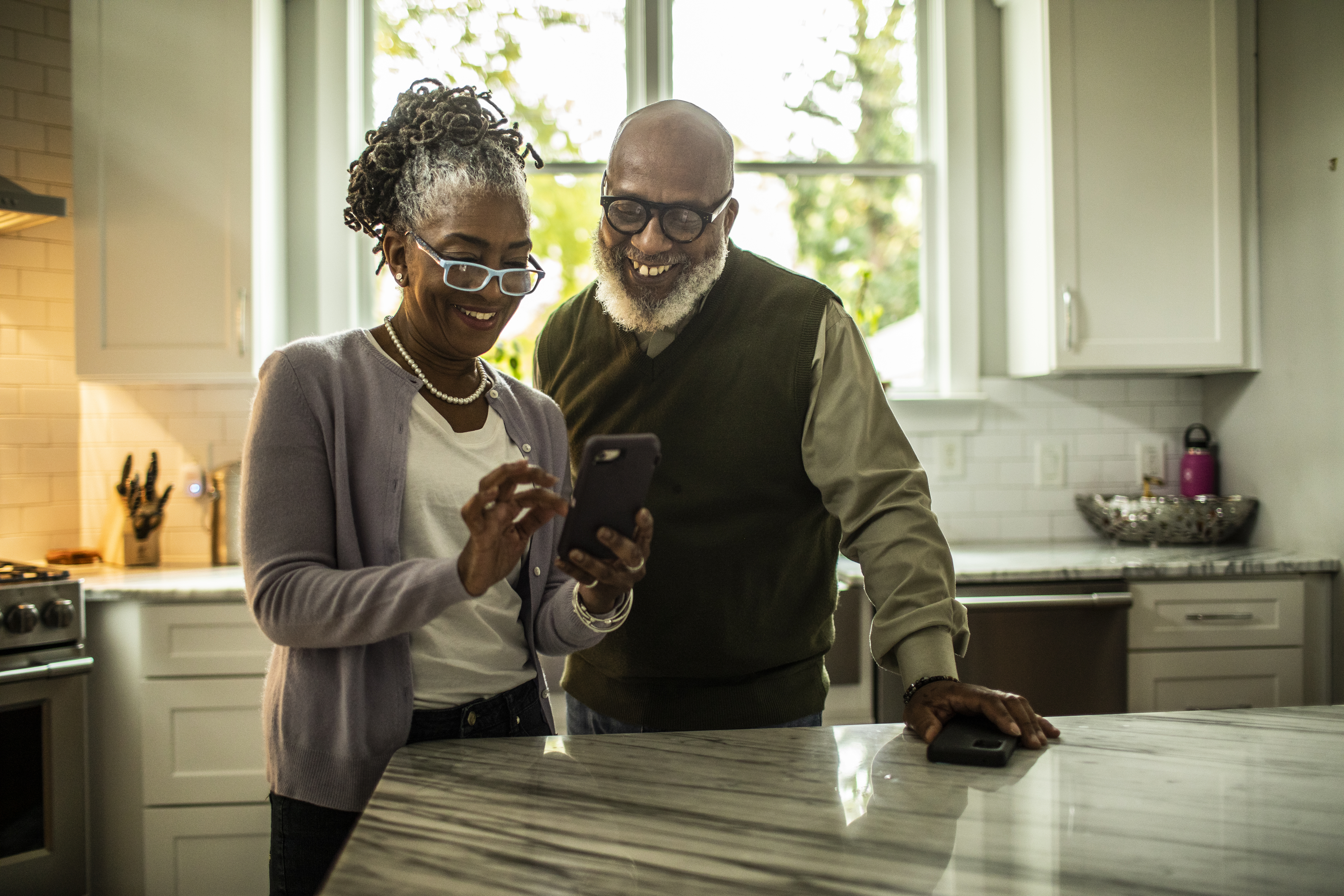 An older couple smiles while looking at a phone together in a bright kitchen