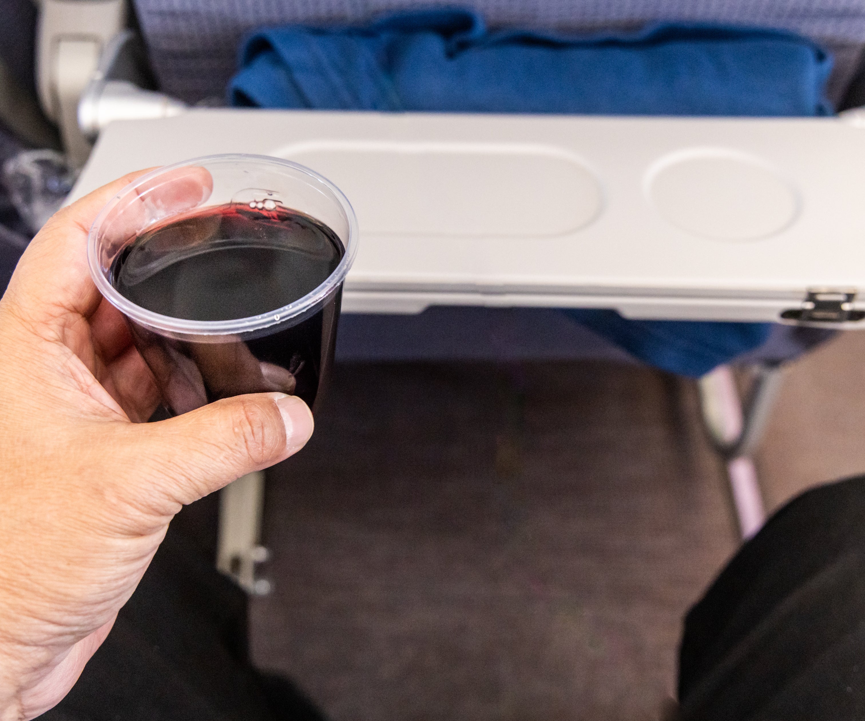 A person holds a plastic cup of red wine while in an airplane seat. The tray table in front is folded down