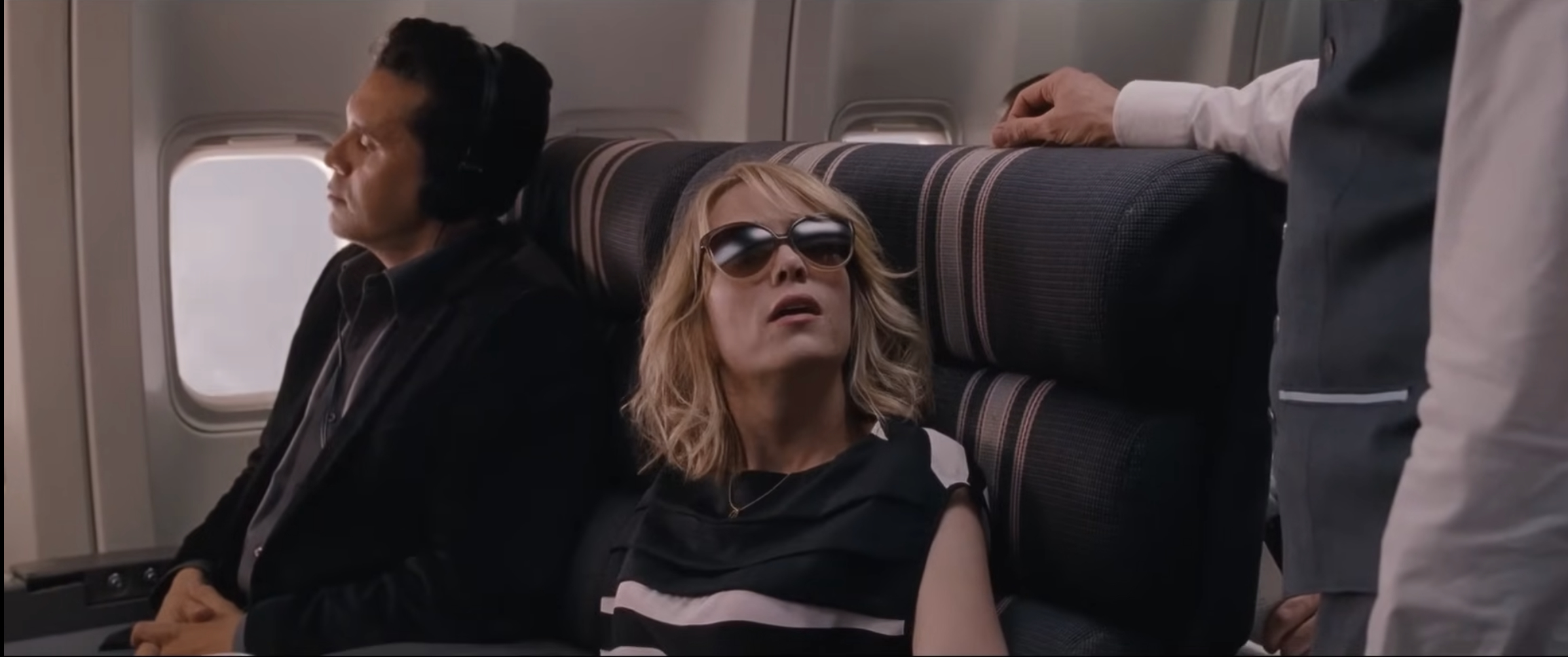 Kristen Wiig sits in an airplane seat wearing sunglasses, with a man in a suit next to her and a flight attendant standing by