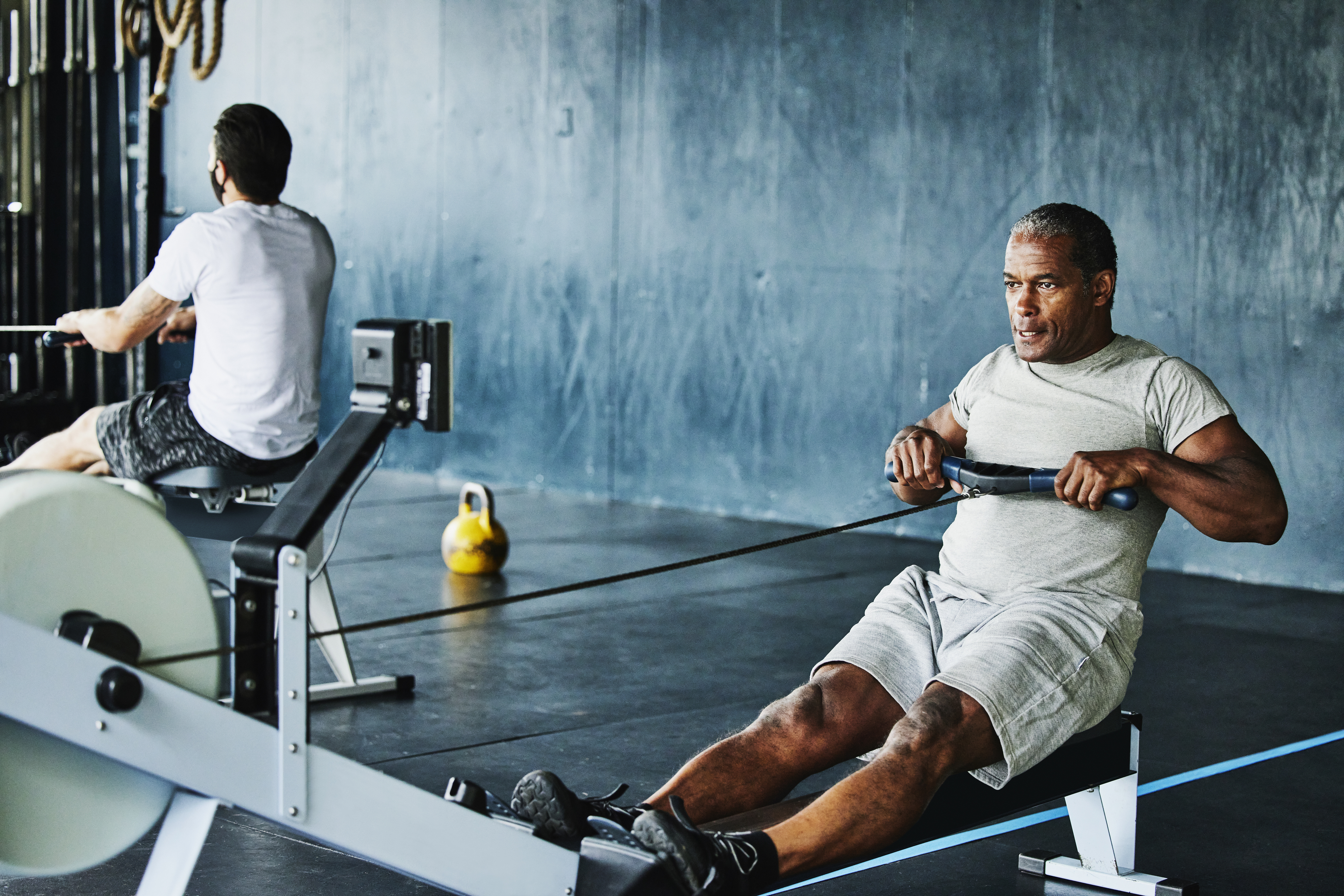 Two men exercising on rowing machines in a gym; one is in the foreground, and the other is in the background