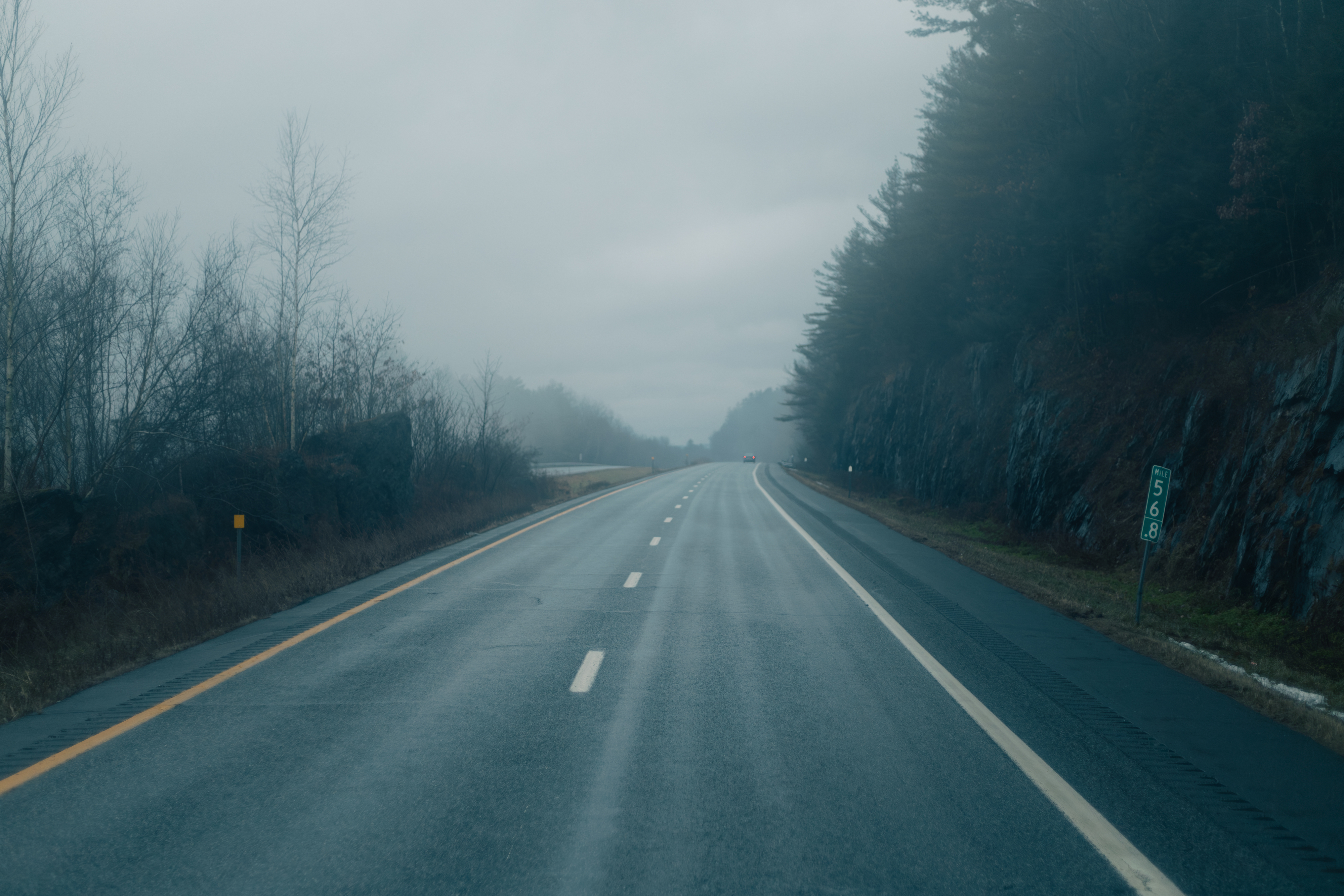 Foggy, empty highway with trees and rocky cliffs on either side, stretching into the distance. No people or vehicles present