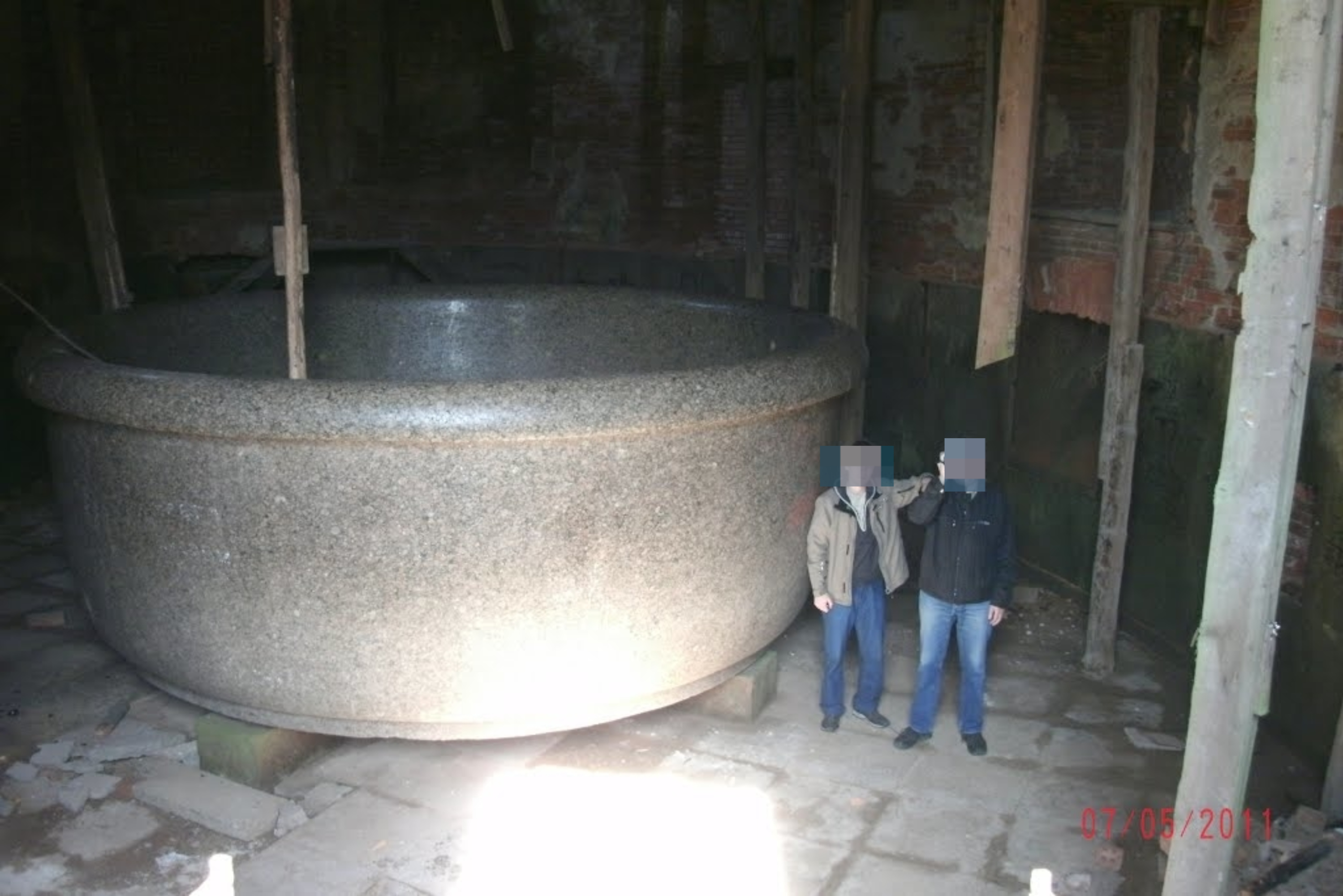 Two people stand next to a massive, circular granite bathtub in a partially dilapidated building. The image date reads 27-08-2011