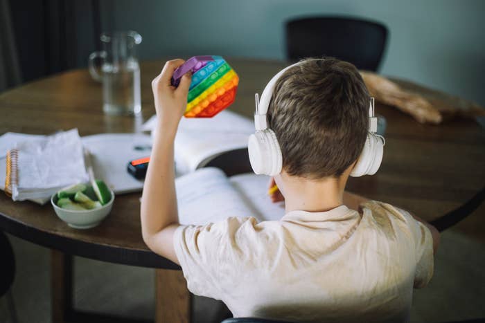 Child wearing headphones holds a rainbow-colored pop-it toy while sitting at a table with an open book, papers, and a bowl of fruit