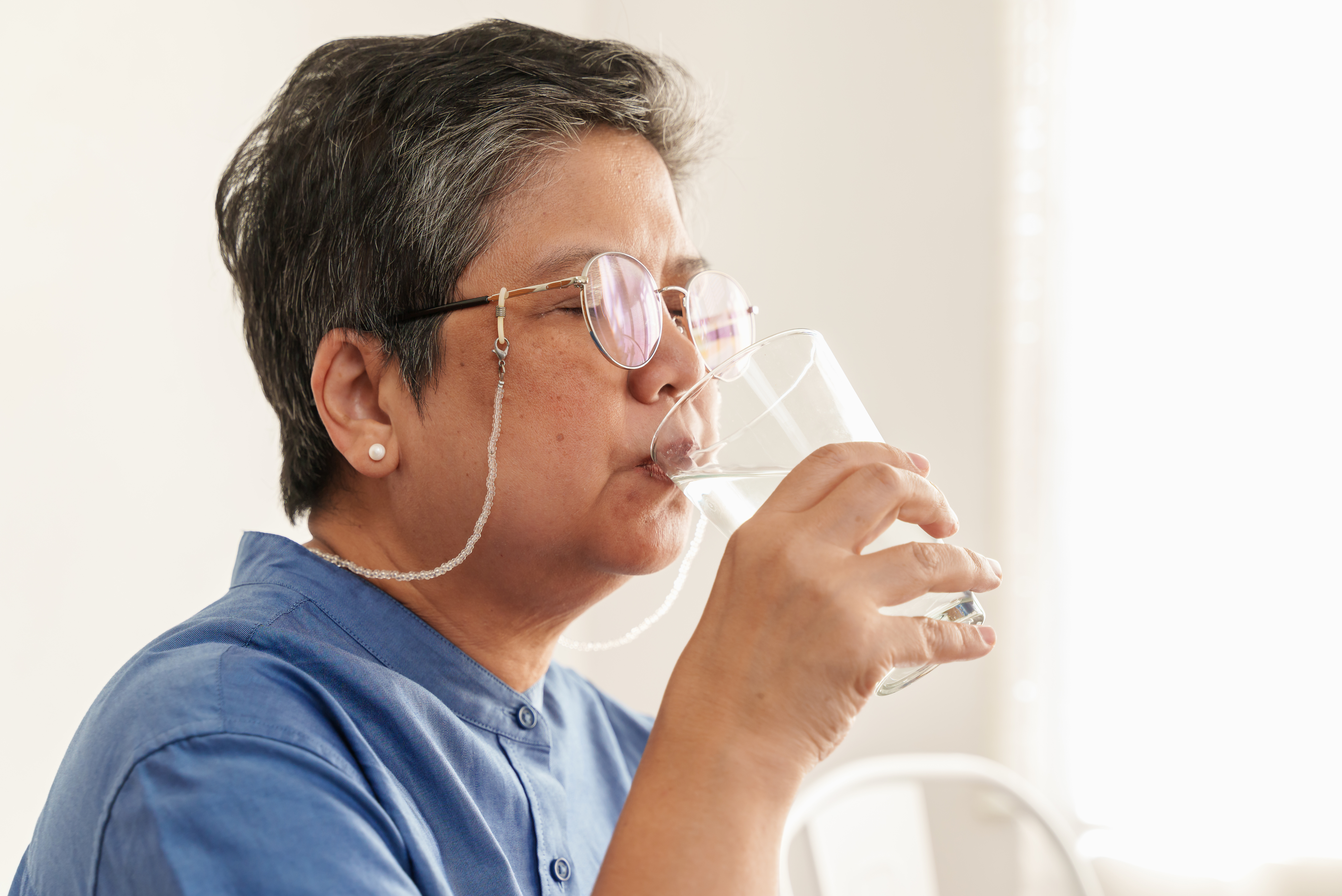 Person wearing glasses and a blue shirt drinks a glass of water with a relaxed expression