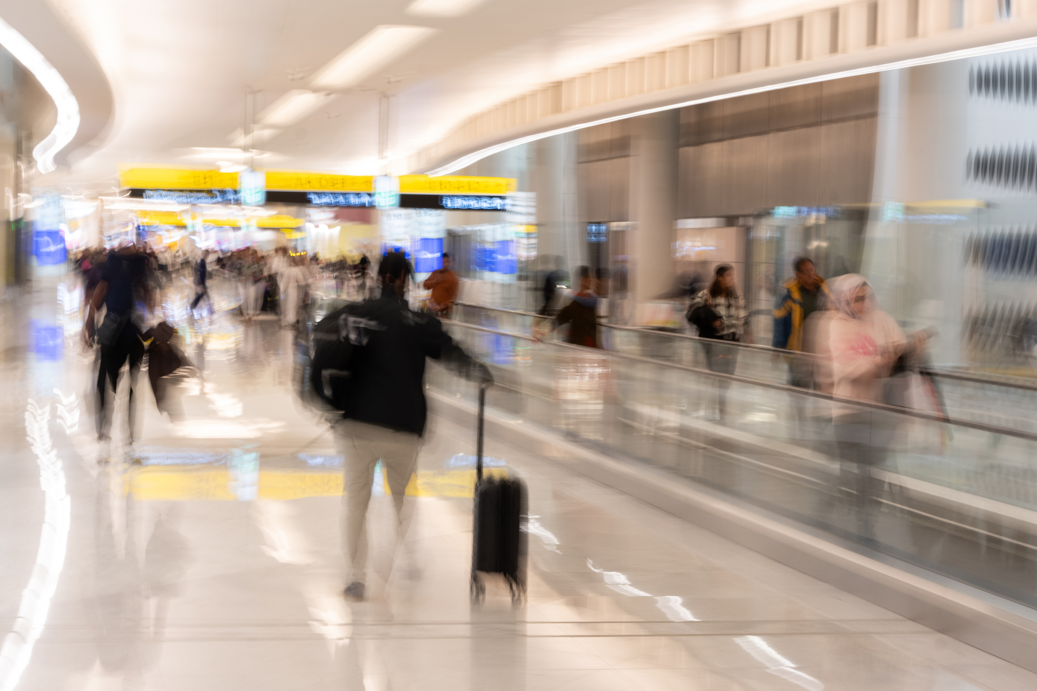 A busy airport terminal with travelers walking and using moving walkways, some with luggage. The image conveys movement and the hustle and bustle of an airport environment