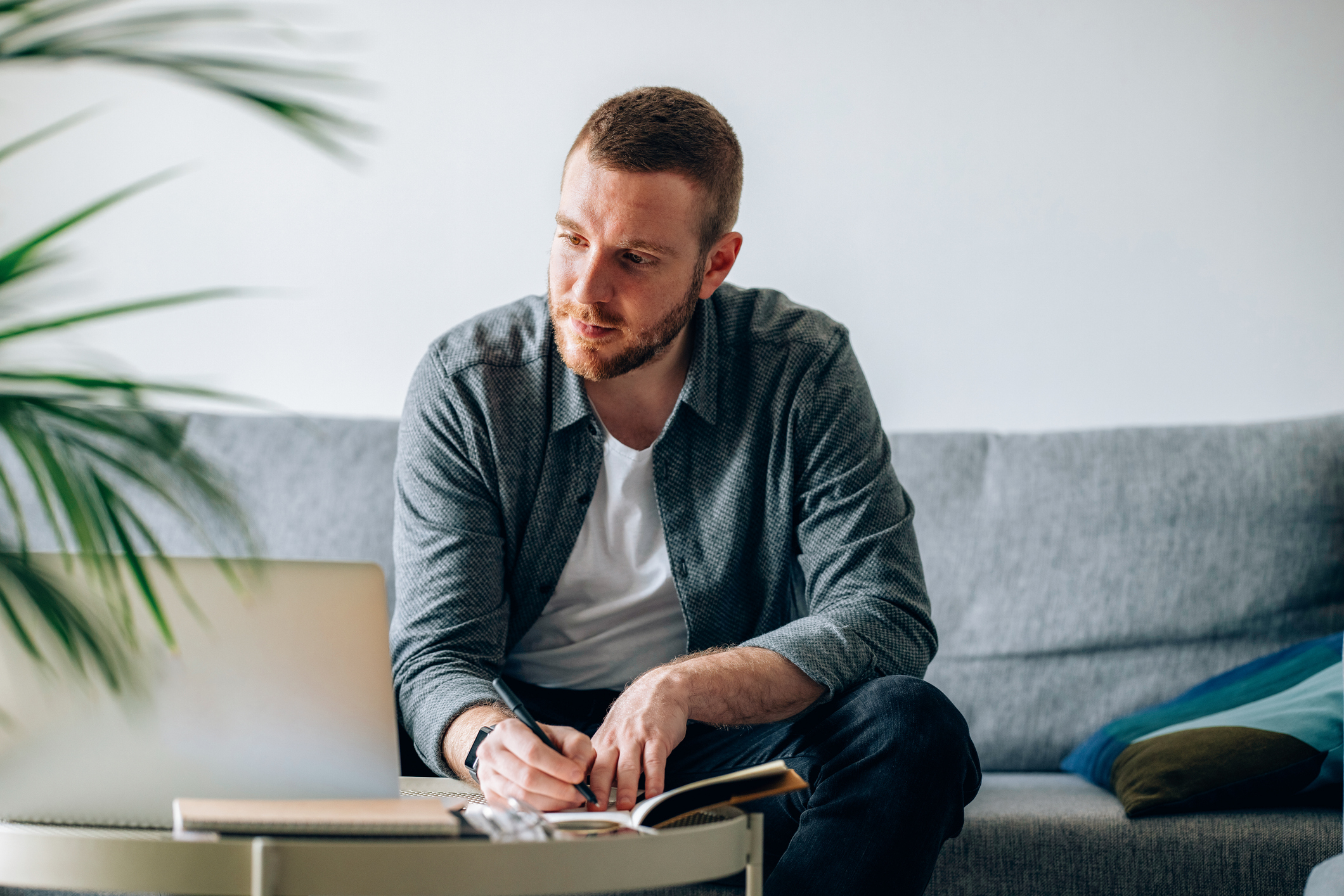 A man sits on a couch, writing in a notebook, while looking at a laptop. He appears focused and engaged in his work