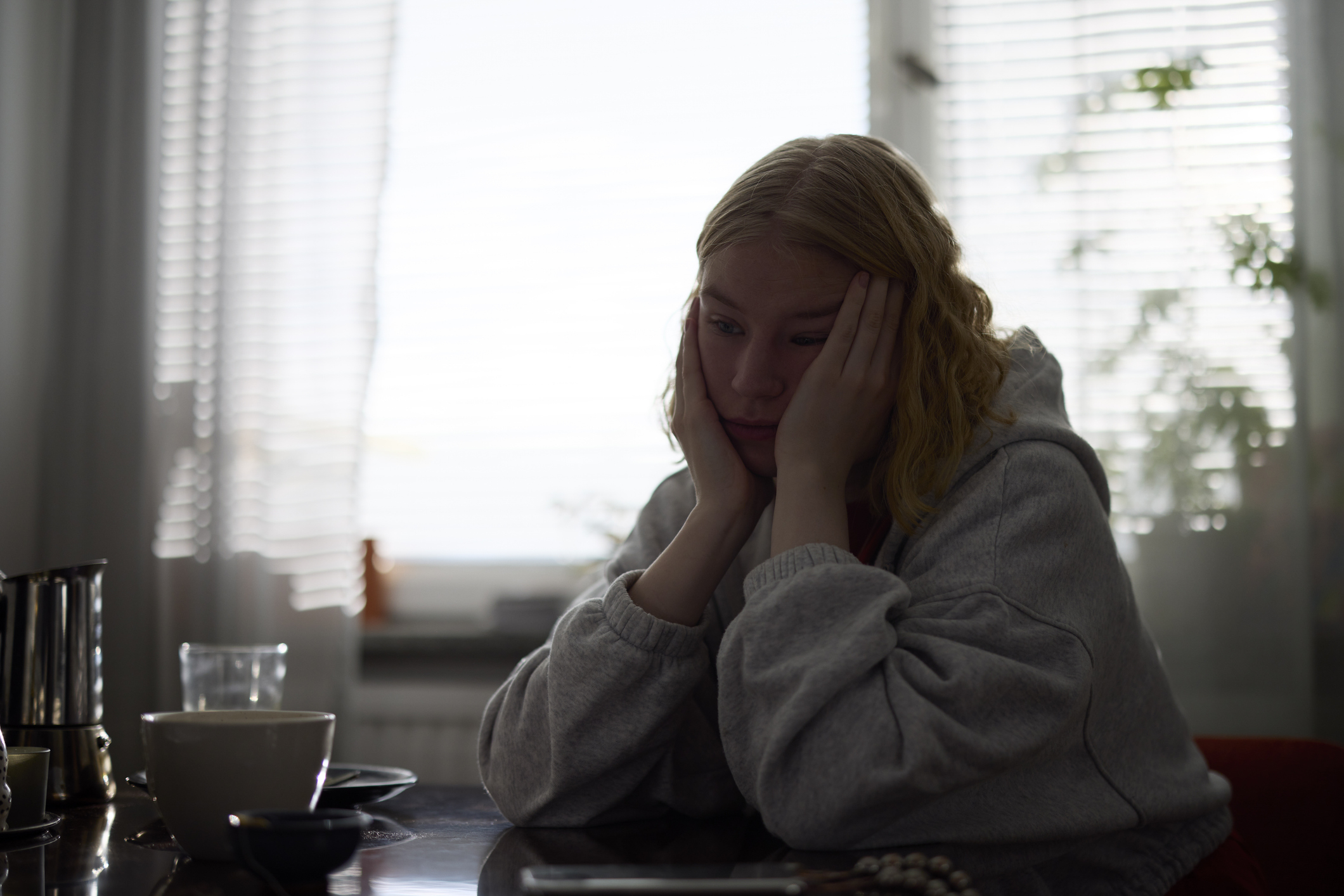 A person with blonde hair, wearing a hoodie, sits at a table with their head in their hands, appearing contemplative and pensive, surrounded by household items