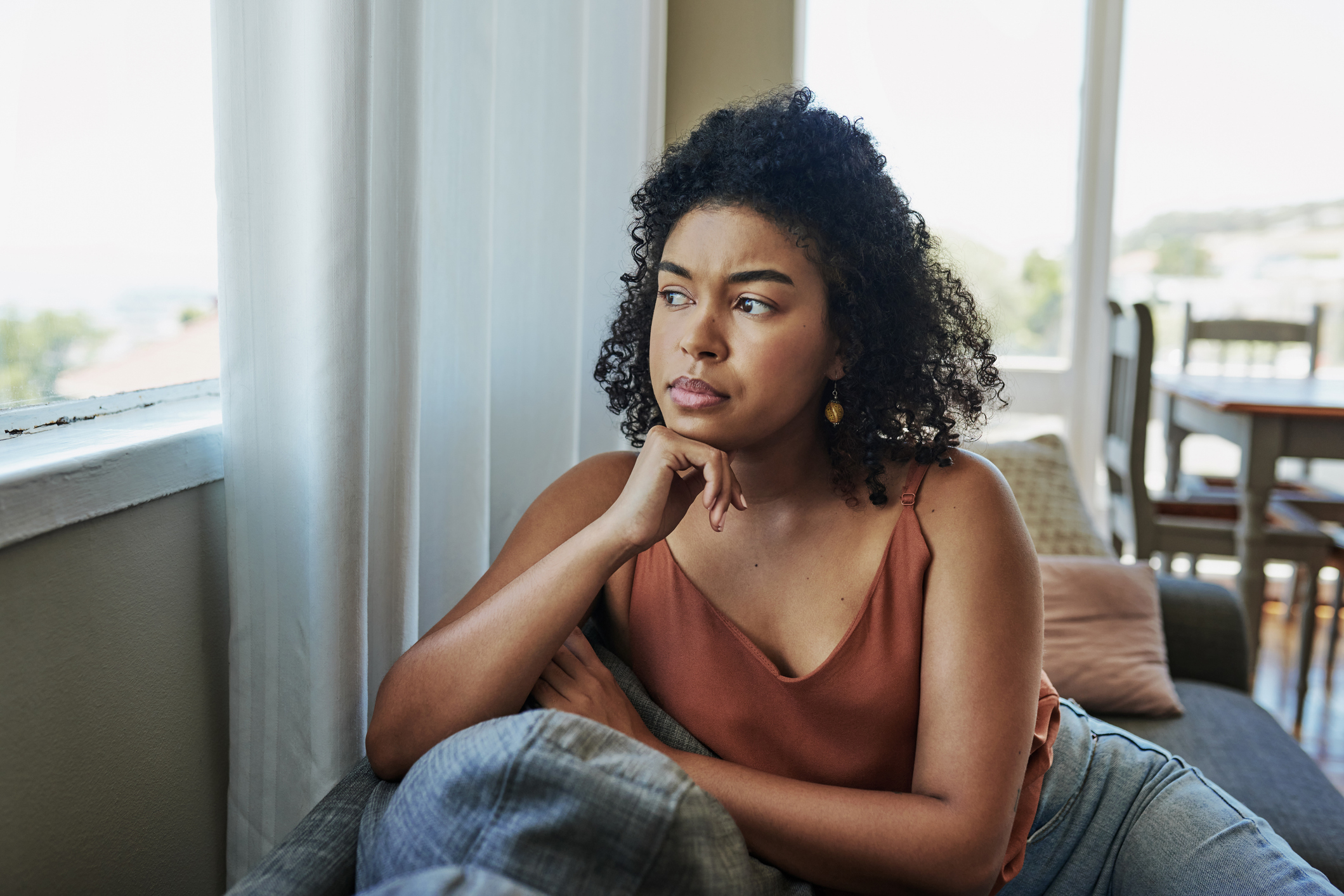A woman with curly hair, wearing a sleeveless top, sits on a couch, leaning on her hand, looking contemplatively out a window