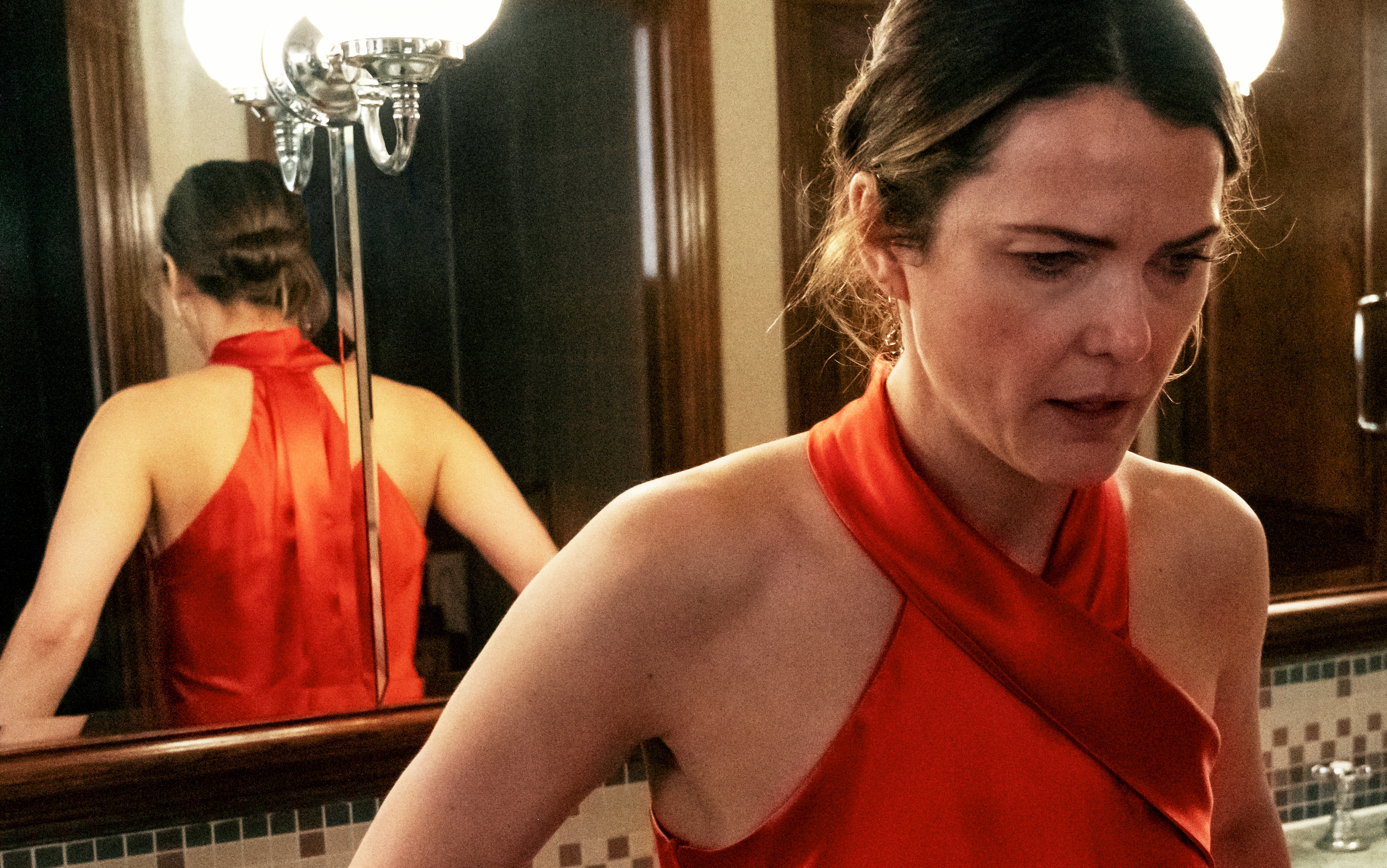 Keri Russell is in a halter-neck dress, seen standing in front of a bathroom mirror with a concerned expression. Her reflection is also visible