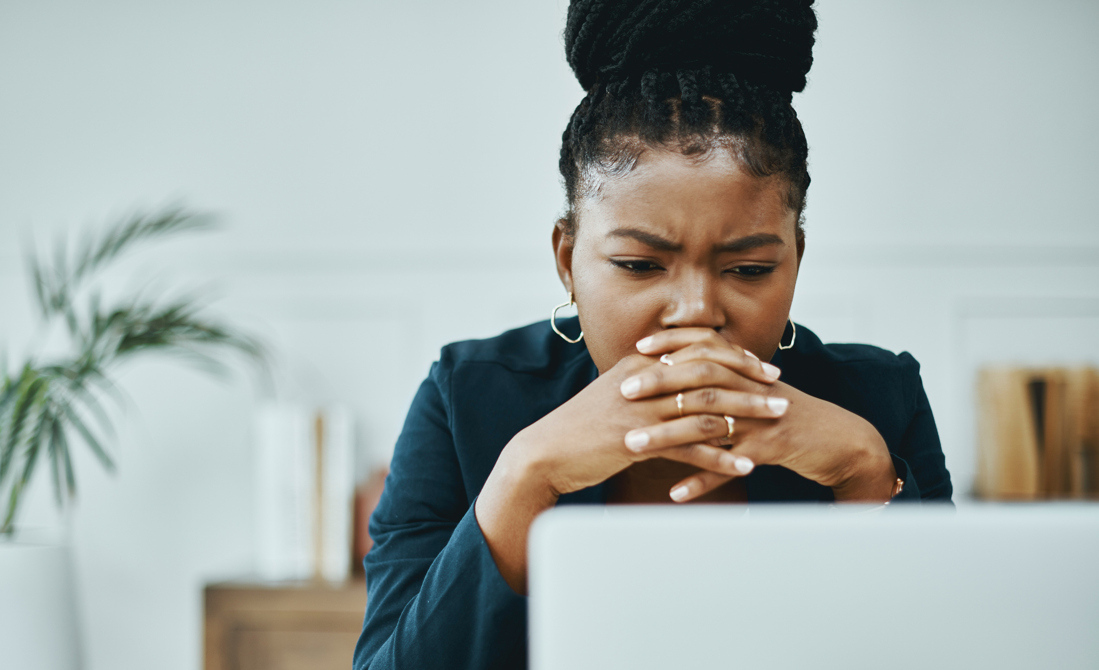 Woman with braided hair and hands clasped in front of her face, looking intently at a laptop screen. Plant and bookshelves in the background