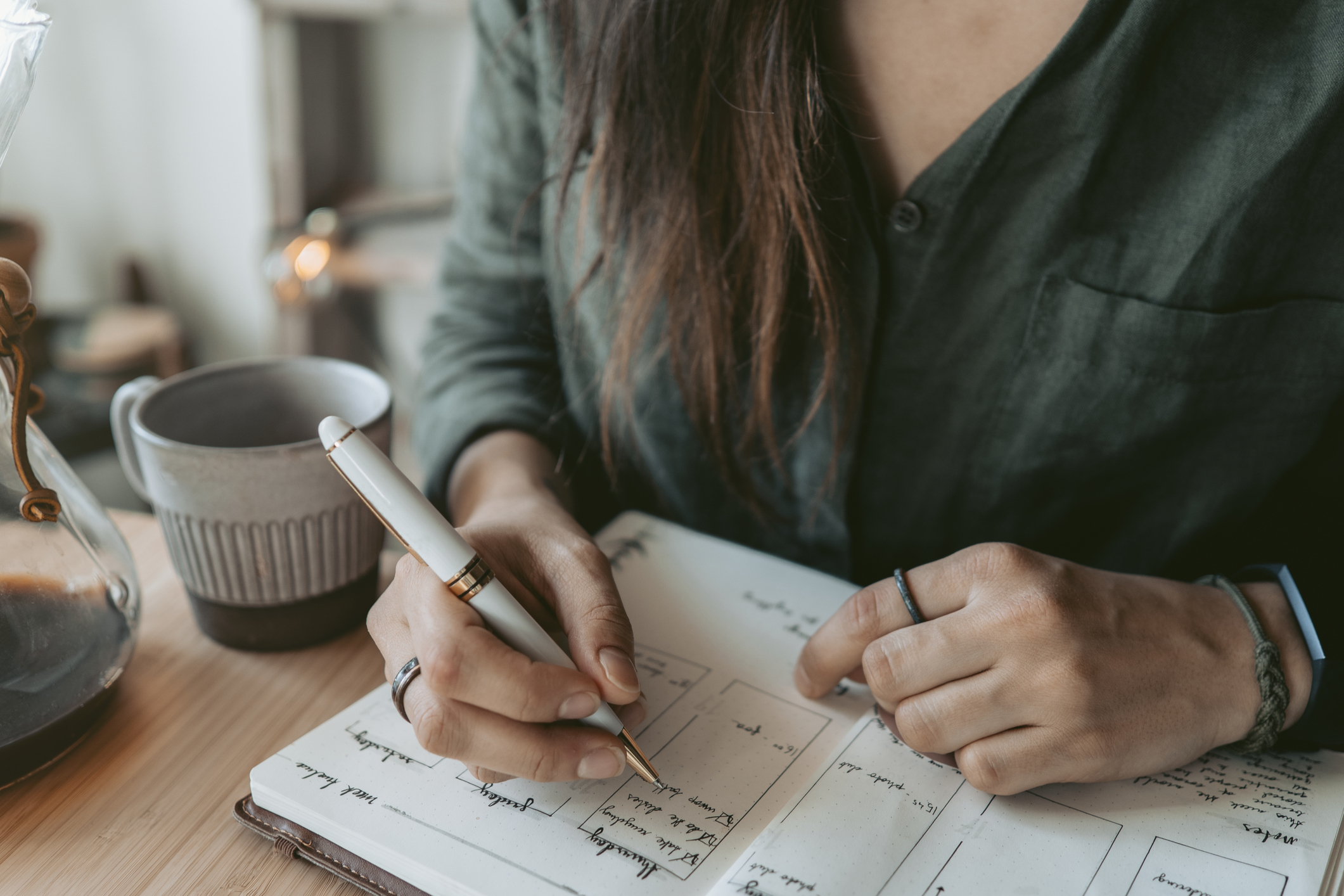 A person writes in a planner at a wooden table. A cup of coffee and a carafe are nearby