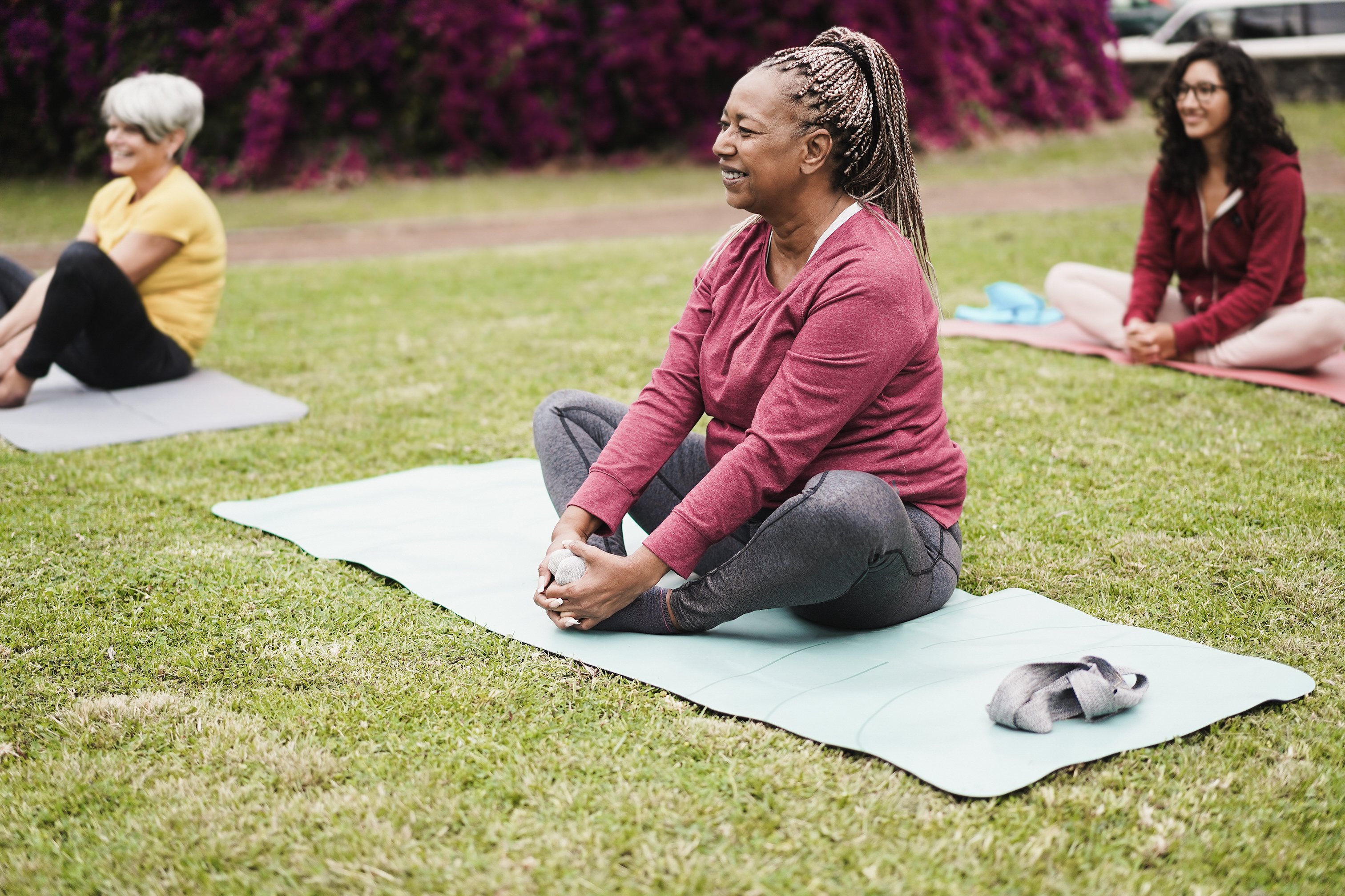 Three people, including an older woman in a red top and gray pants, perform seated yoga poses on mats outside in a park