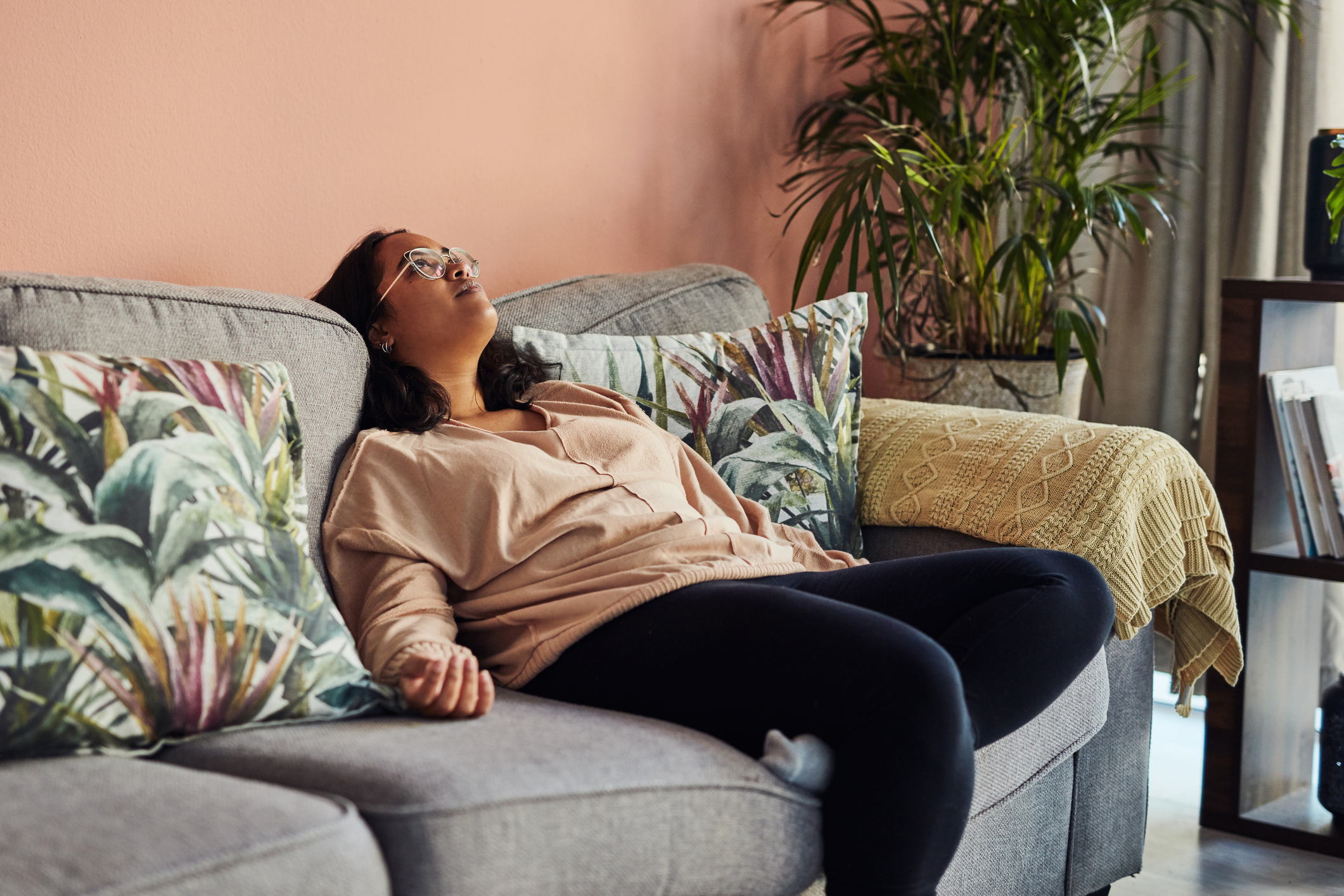 Woman sits on a couch, leaning back with a relaxed expression, in a cozy living room with plant-themed pillows and potted plants