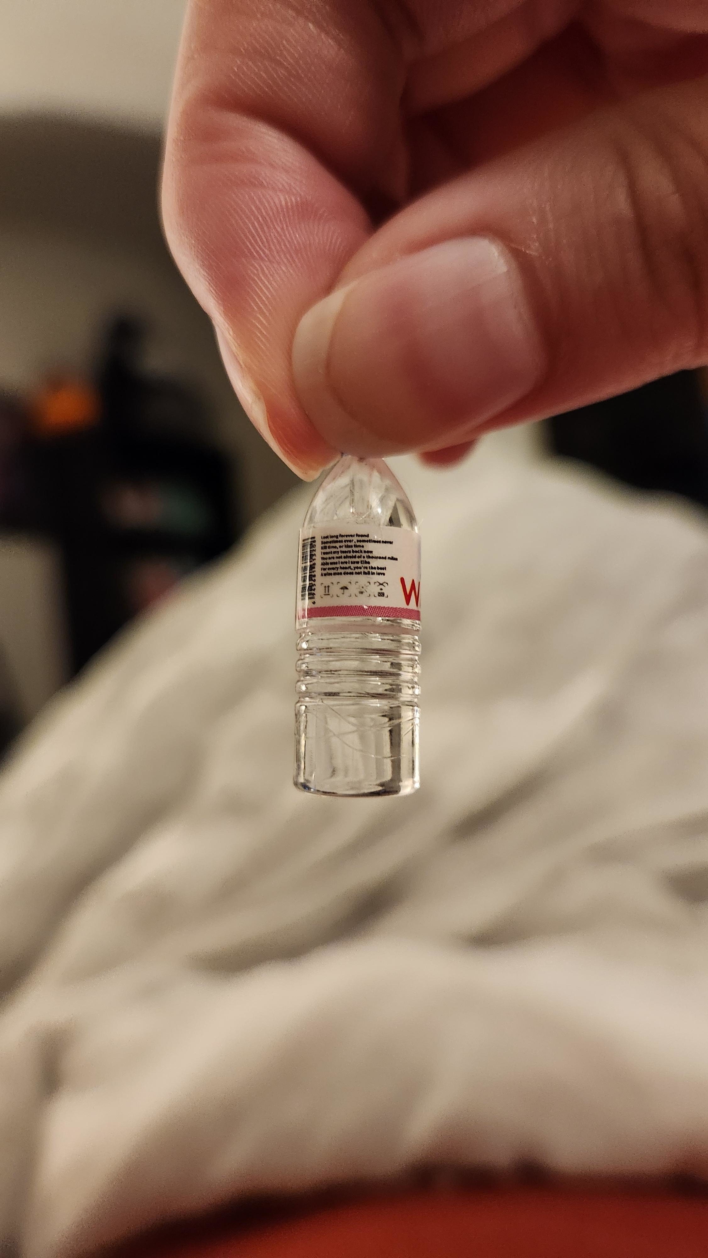 A hand holding a tiny plastic bottle resembling a water bottle. Background includes a blurred indoor setting with what appears to be a bed