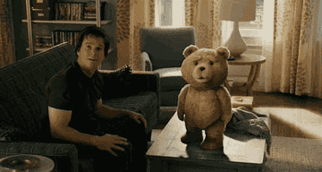 Mark Wahlberg sits on a couch next to a talking teddy bear named Ted, who is standing on a coffee table in a living room setting