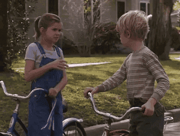 Two children, a girl in overalls and a boy in a striped shirt, stand with bikes on a residential street