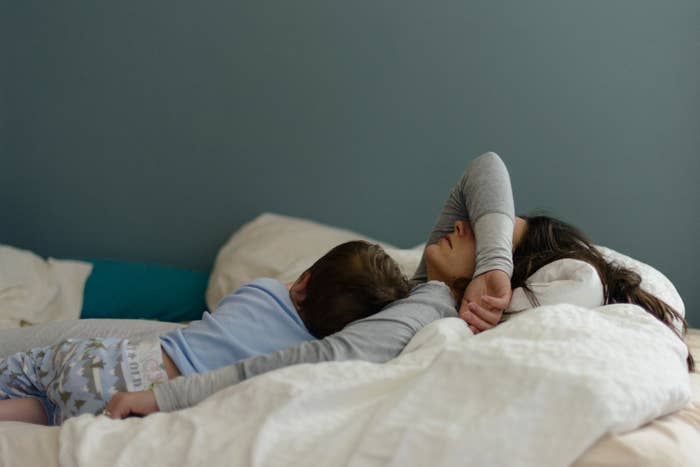 A woman is lying in bed with her arm over her face, and a young child is sleeping next to her, partially on her arm. They both appear to be resting peacefully
