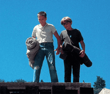 River Phoenix and Corey Feldman stand outdoors holding rolled sleeping bags. River wears a white shirt and jeans, while Corey wears glasses and a dark shirt
