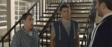 Jonah Hill and Channing Tatum stand in front of a staircase, listening to Rob Riggle, in a scene from "21 Jump Street."