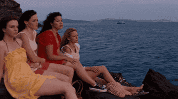 Four friends sit on rocks near the ocean, gazing at the horizon, in casual summer outfits
