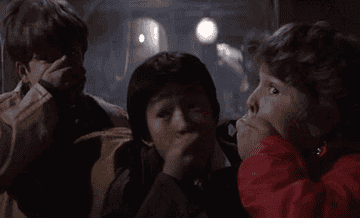 Three children from The Goonies stand closely, covering their mouths in surprise. Names unknown. The background is dark, indicating a nighttime or indoor scene