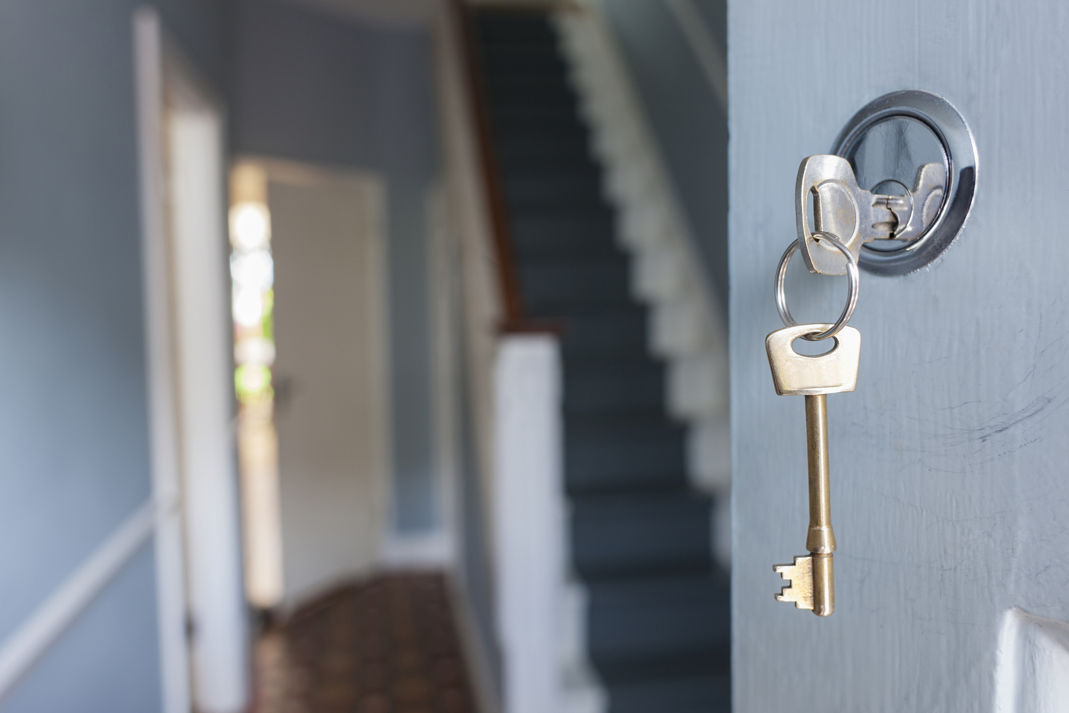 Close-up of keys hanging in a lock inside a home hallway, leading to a staircase. The hallway is softly lit with an open door in the background