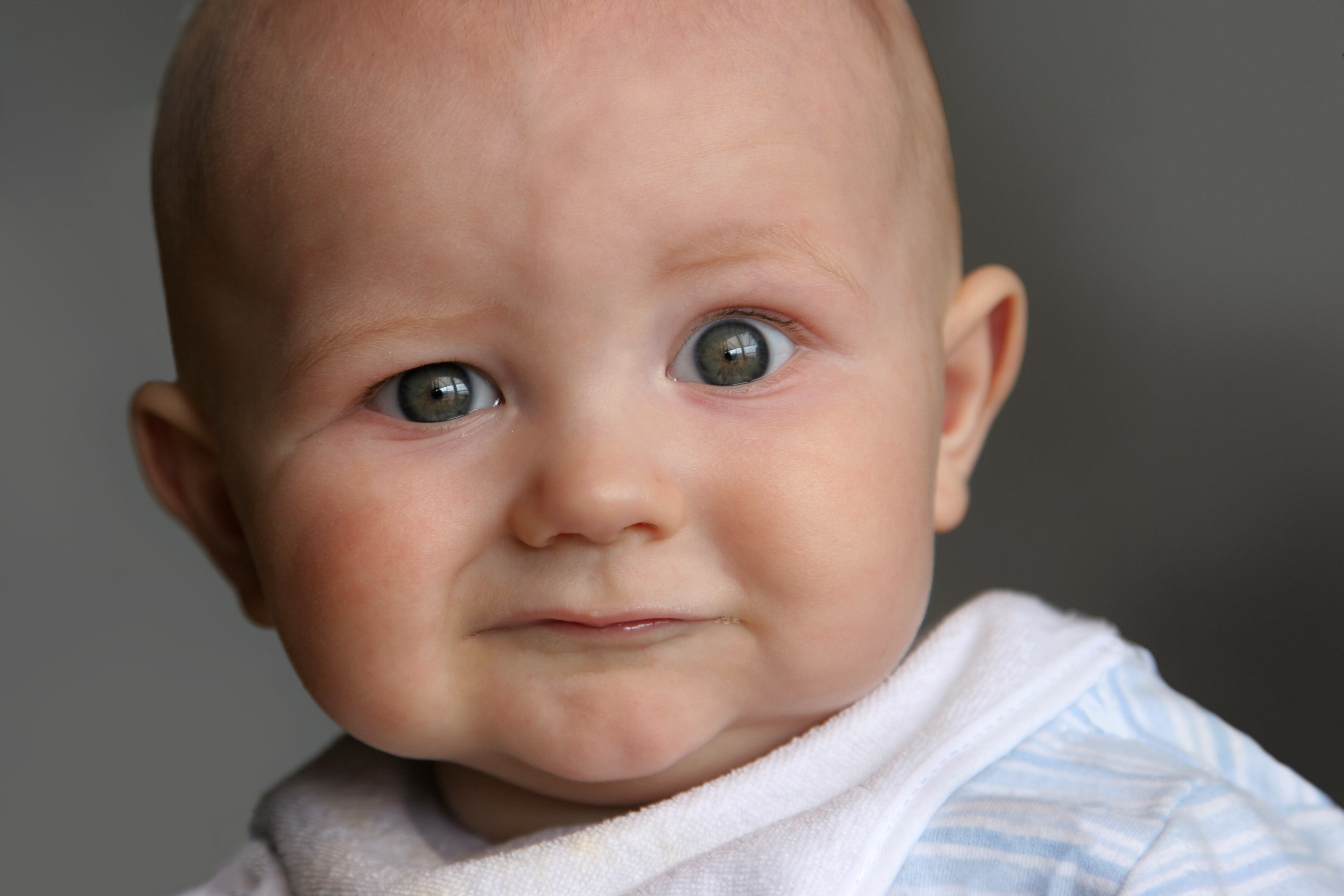 A close-up of a baby wearing a bib and a patterned onesie, looking directly at the camera with a curious expression