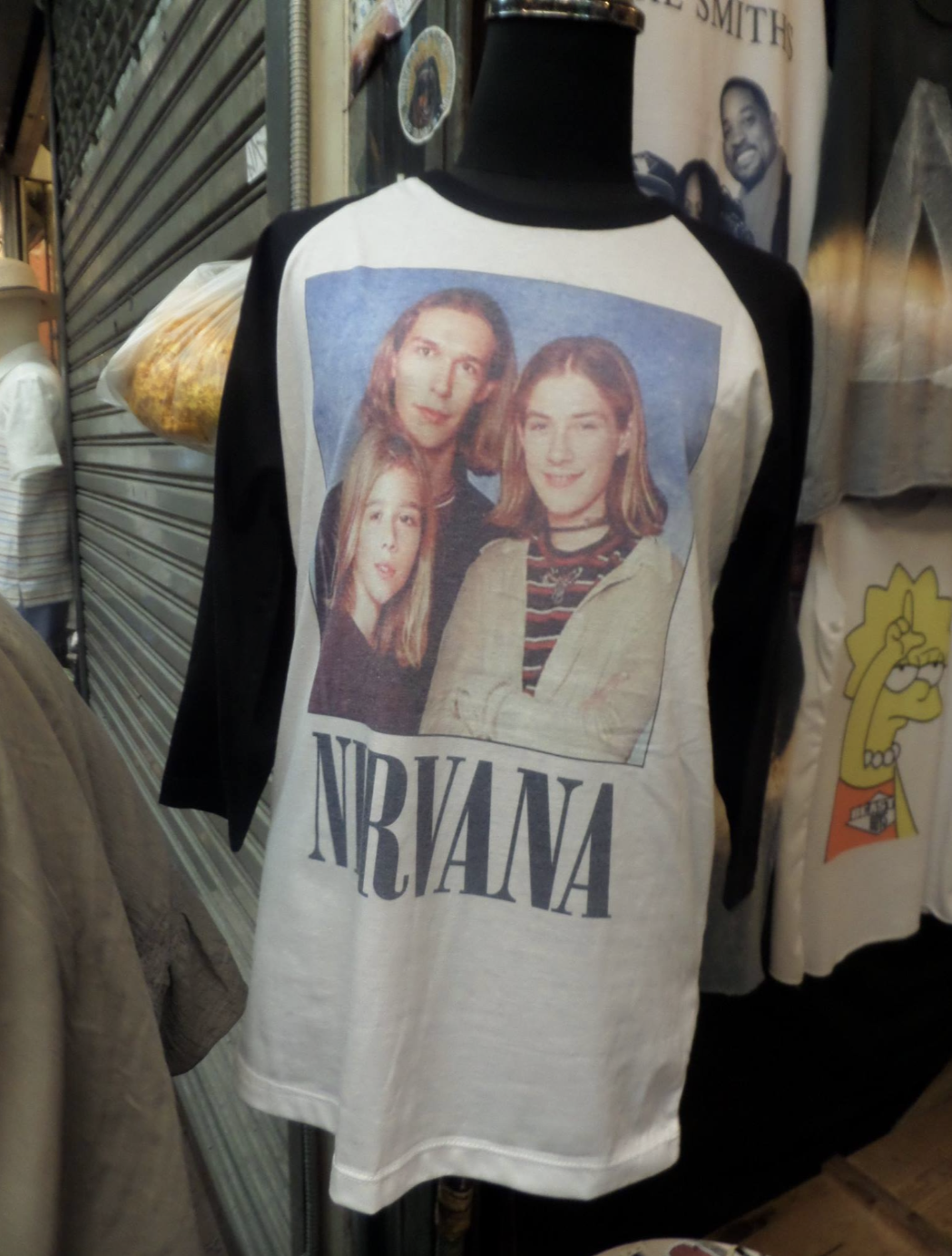 Mannequin wearing a shirt with "Nirvana" text and an image of three smiling individuals with shoulder-length hair. Store backdrop with various t-shirts