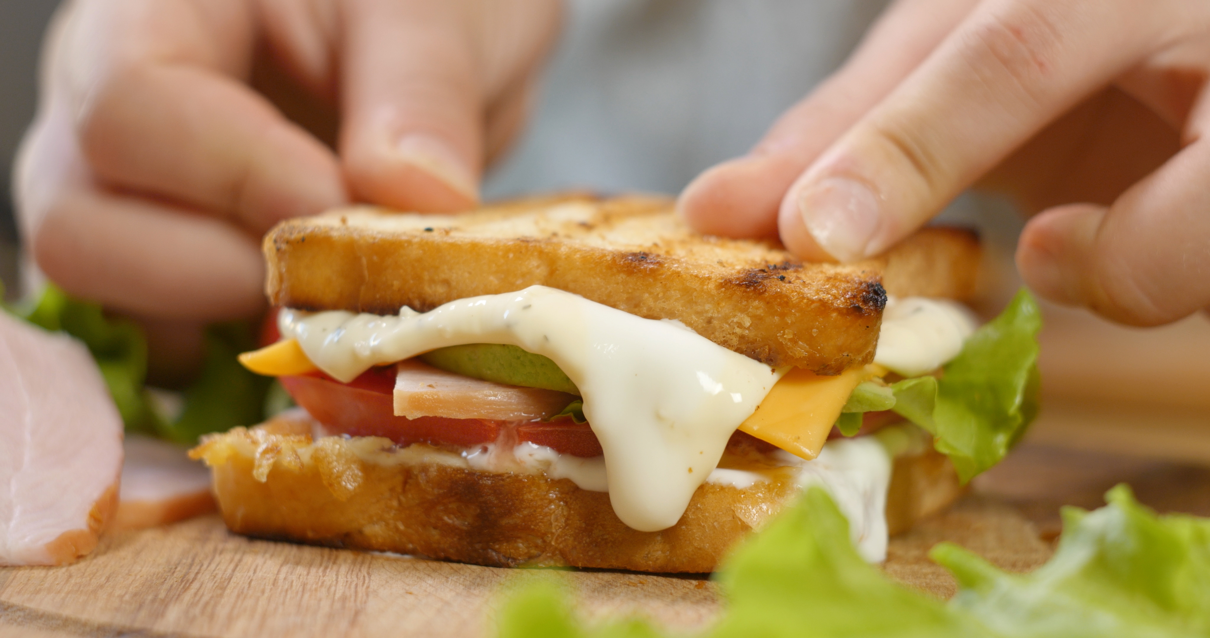 Close-up of a grilled sandwich with lettuce, tomato, cheese, and deli meat being held by two hands. Lettuce and ham slices are in the background