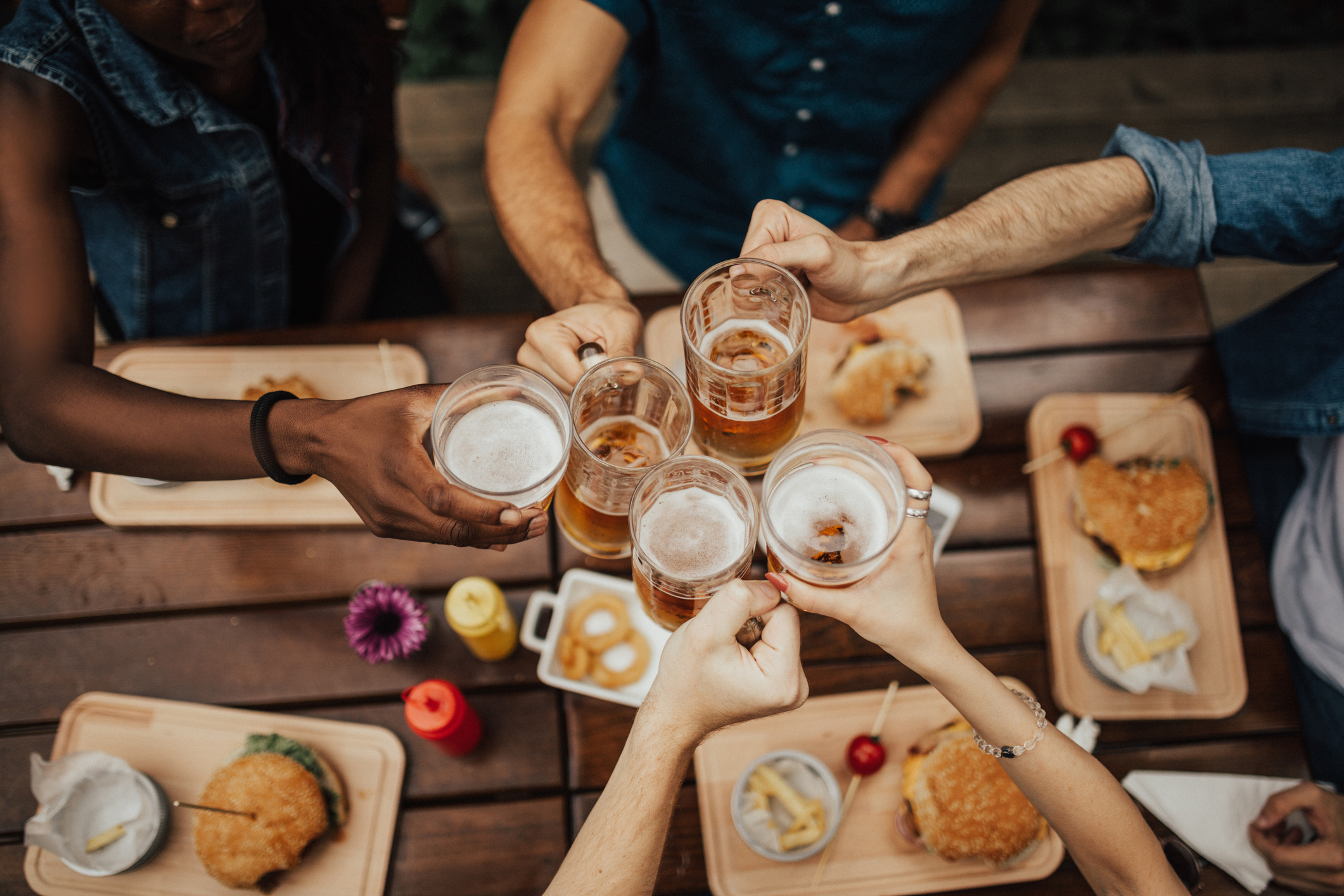 People raising their beer mugs for a toast over a table with burgers and fries on wooden trays