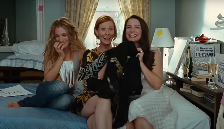 Sarah Jessica Parker, Cynthia Nixon, and Kristin Davis sit on a bed, laughing and holding clothing items, with a cityscape visible through the window behind them