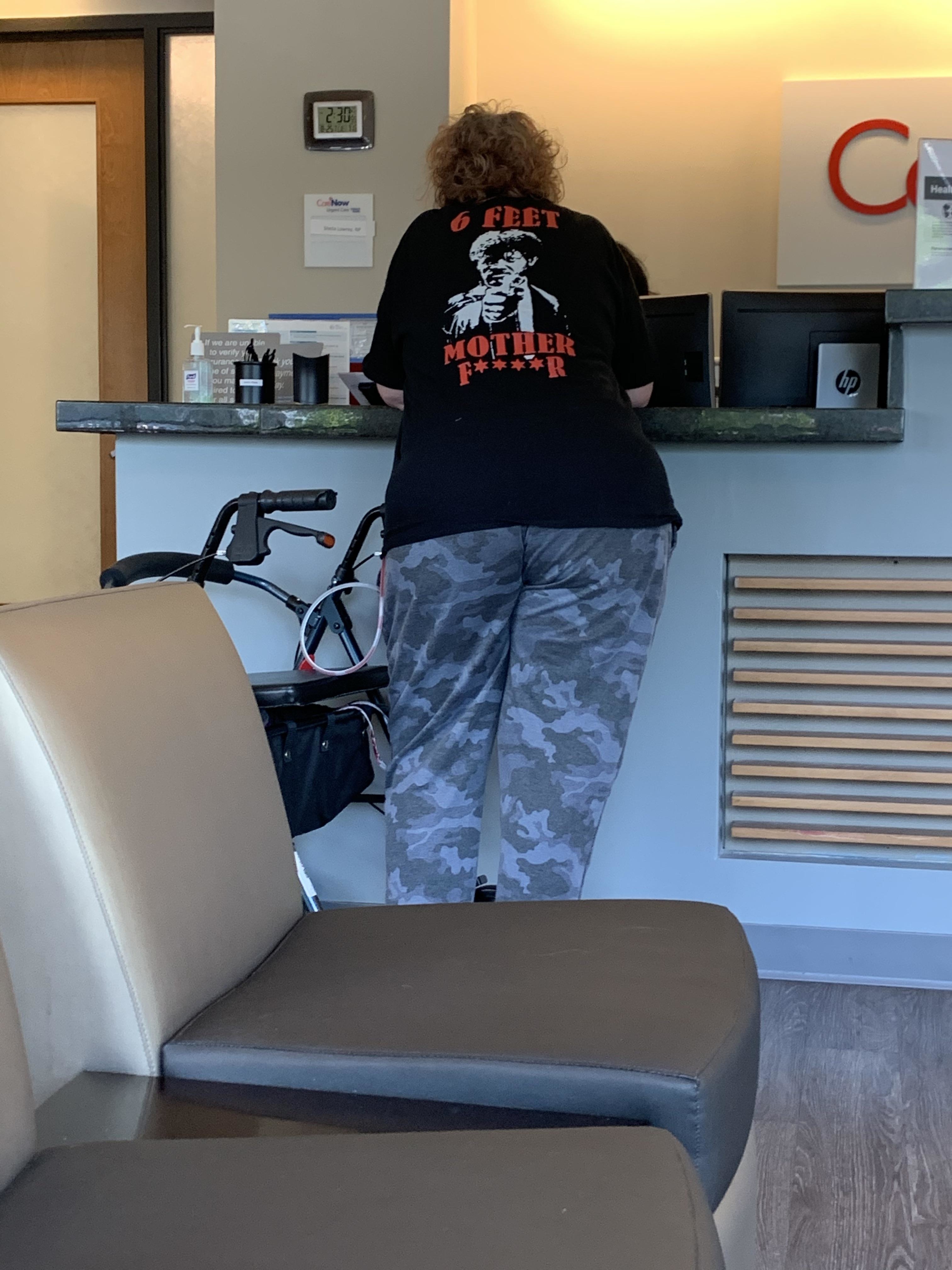 A woman, whose name is unknown, stands at a counter in a public setting. She is wearing a black T-shirt with the text "6 FEET MOTHER F**ER" and camouflage pants