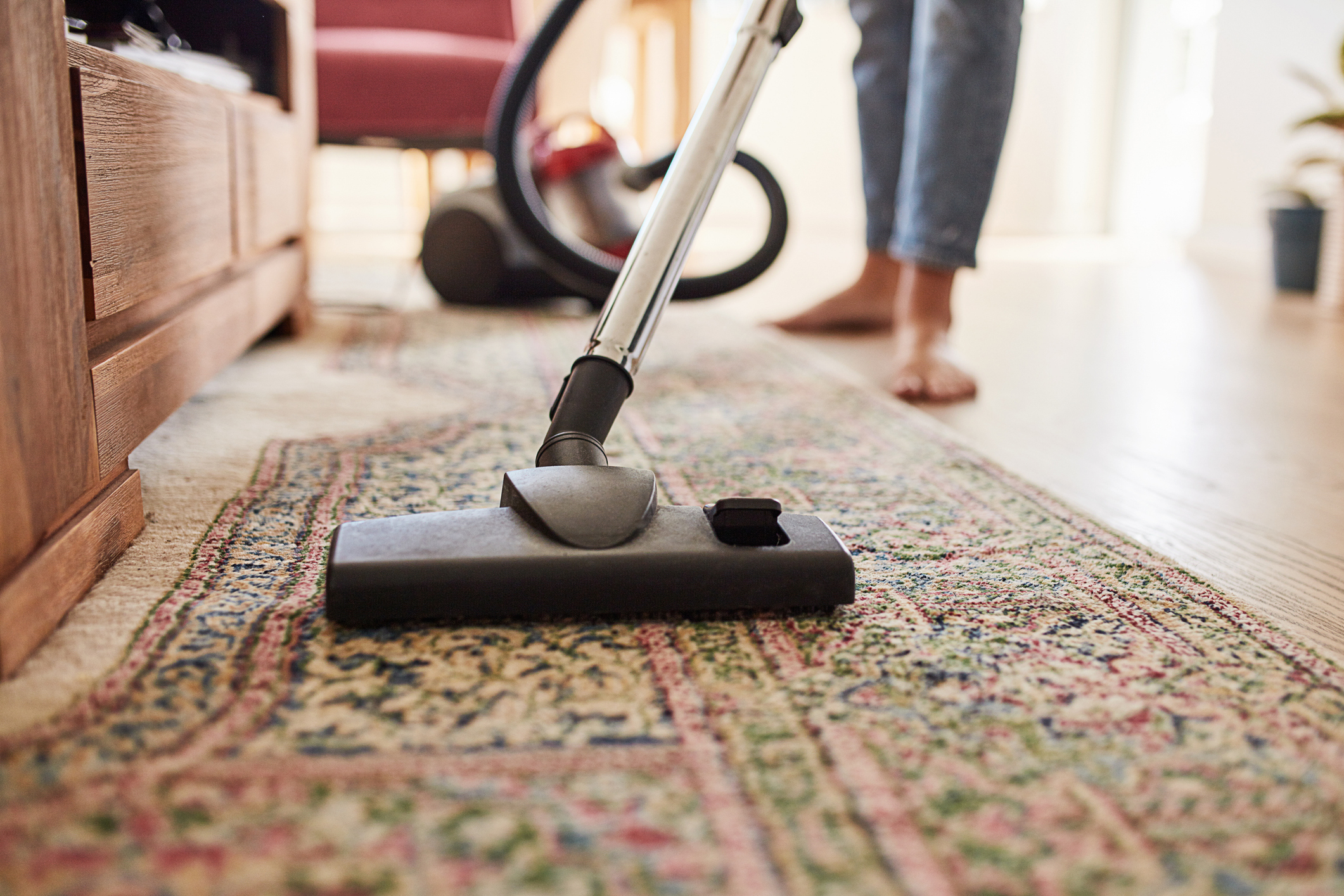 A person vacuuming a patterned rug in a living room setting with part of a red chair and a plant visible in the background