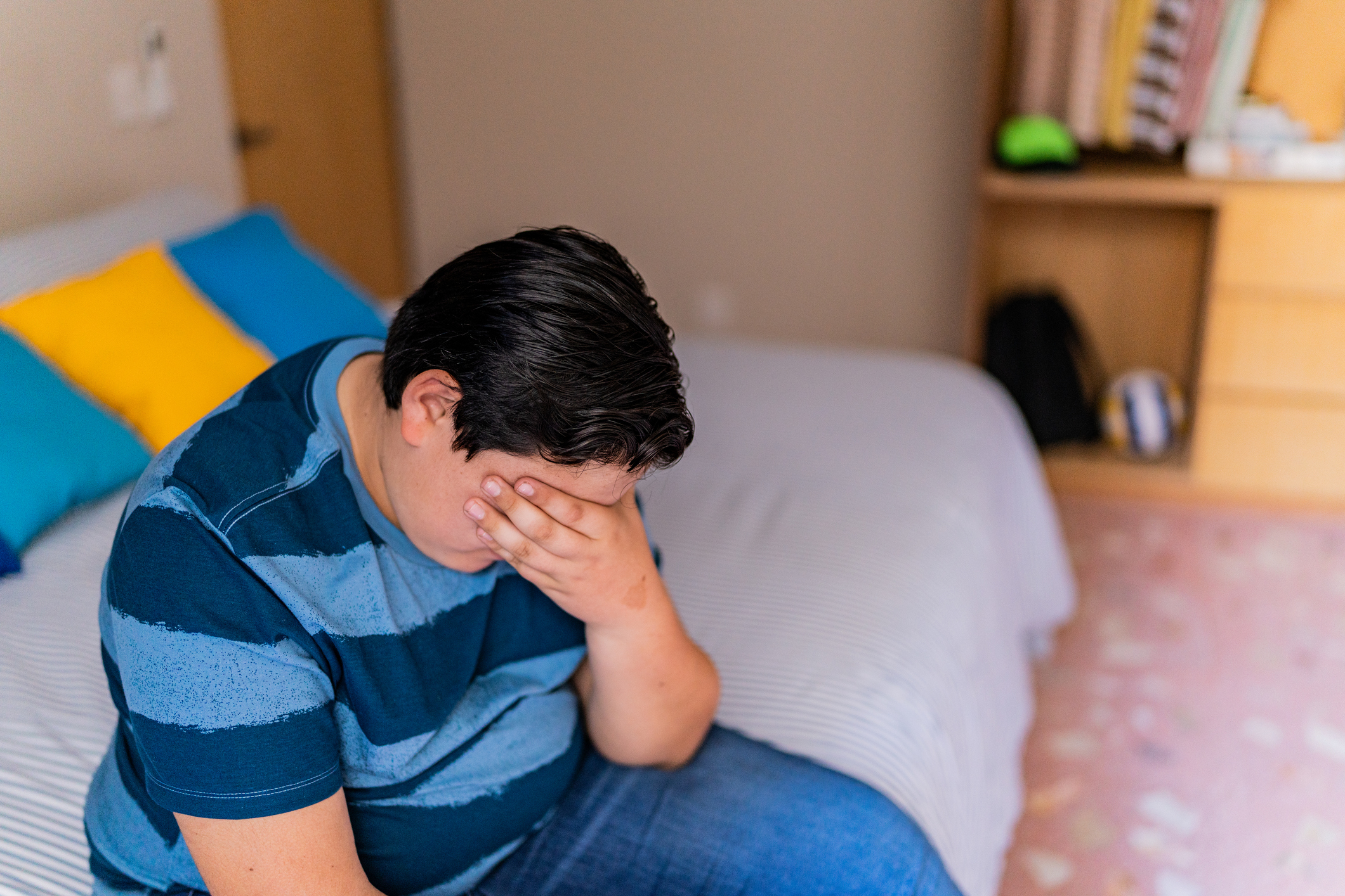 A person, unidentified, sits on a bed appearing stressed with their hand covering their face. The room has cushions and shelves in the background