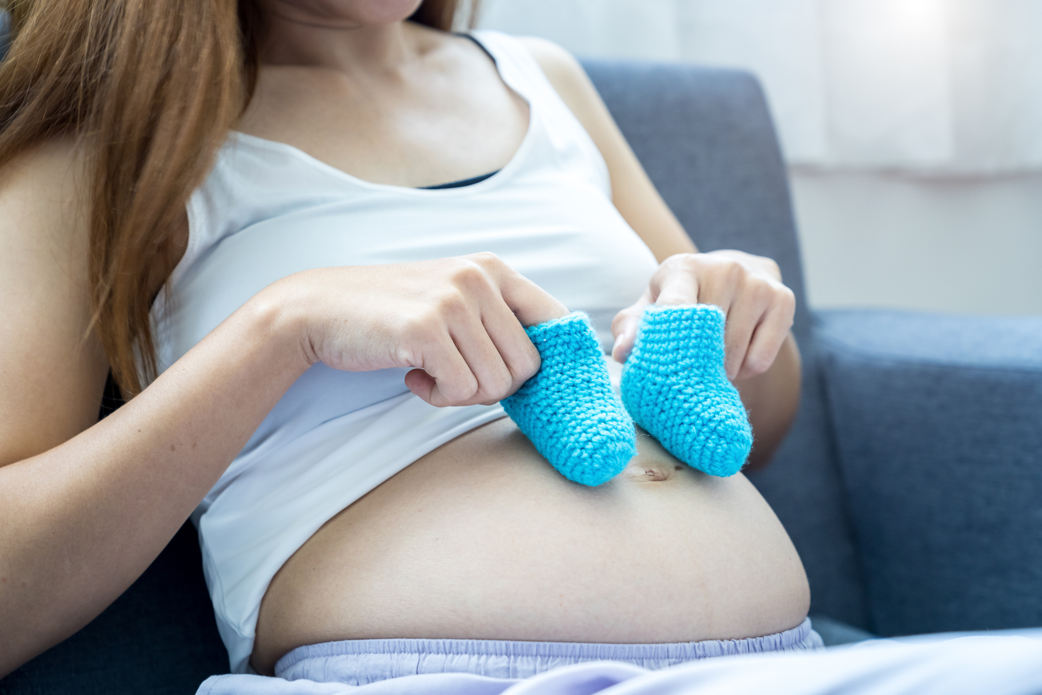 A pregnant woman sits on a couch, holding a pair of small blue baby booties over her bare belly