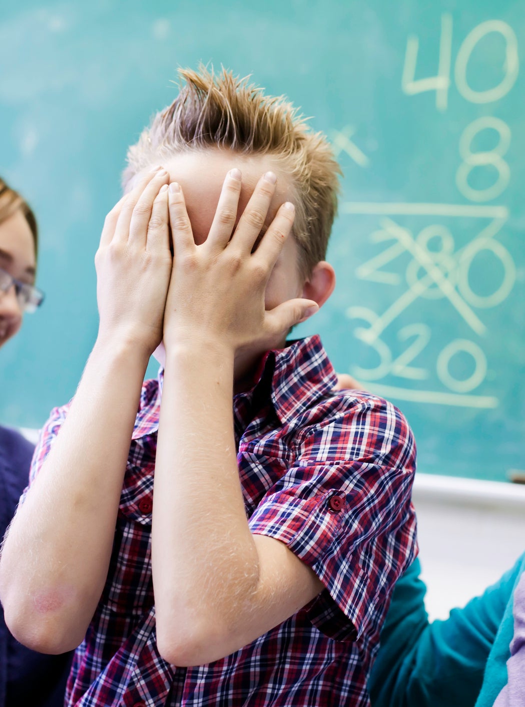 Two children smile at a boy who covers his face with his hands while standing in front of a chalkboard with math problems