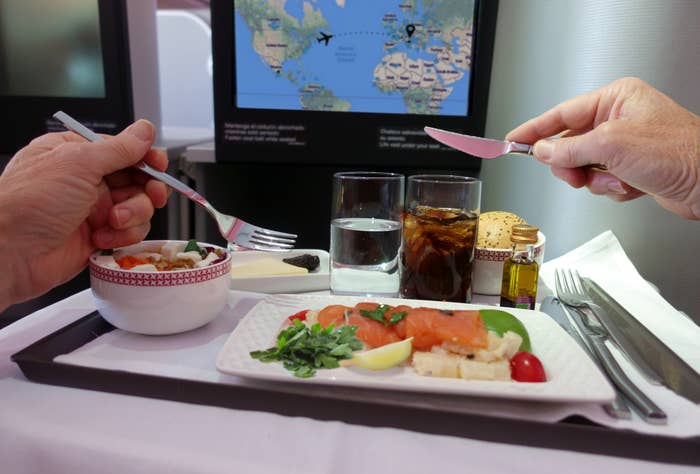 Two people dining on a flight with a meal that includes salmon, vegetables, water, and soda. A world map is displayed on a screen in the background