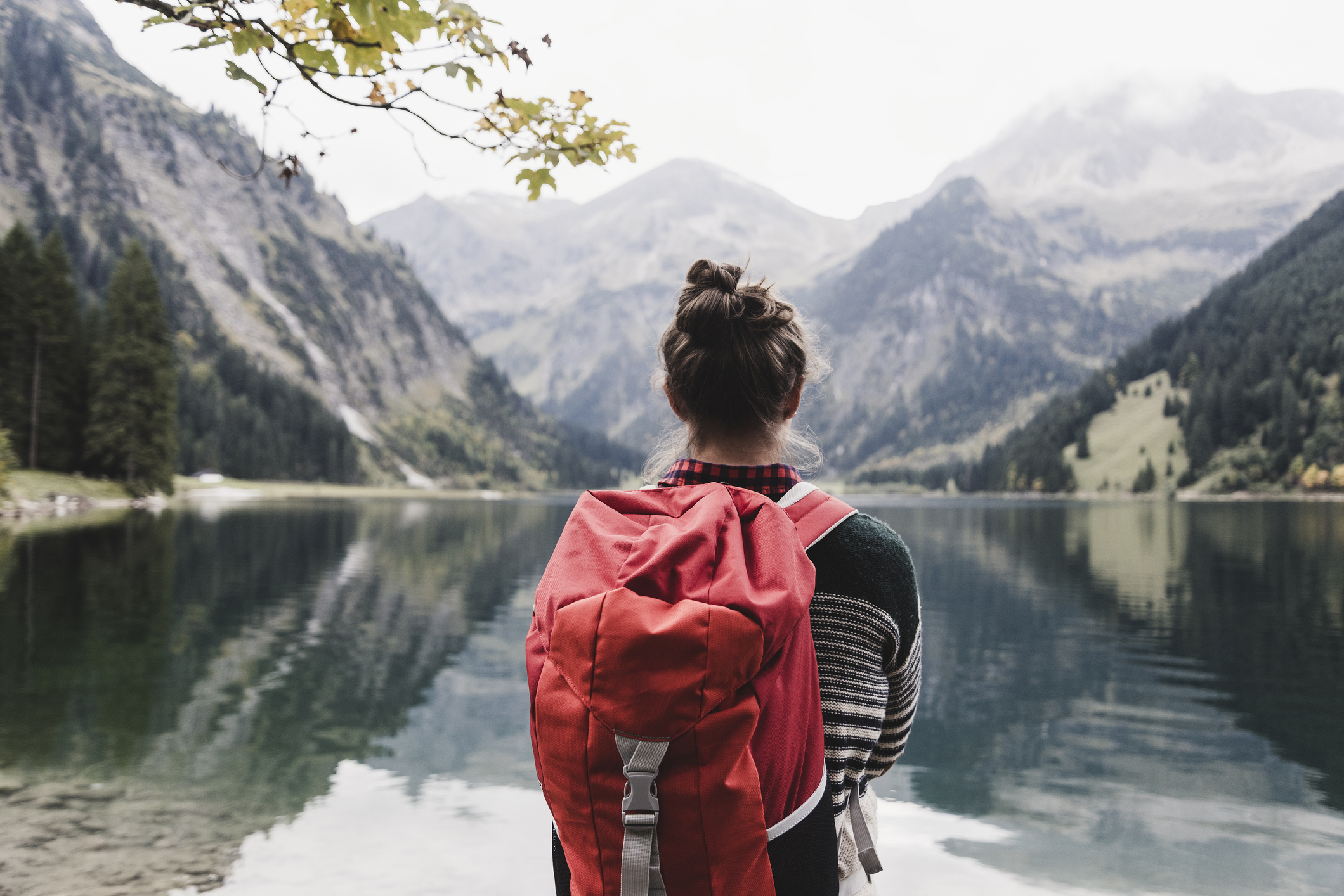 Person with a red backpack looks at a serene lake with mountains in the background