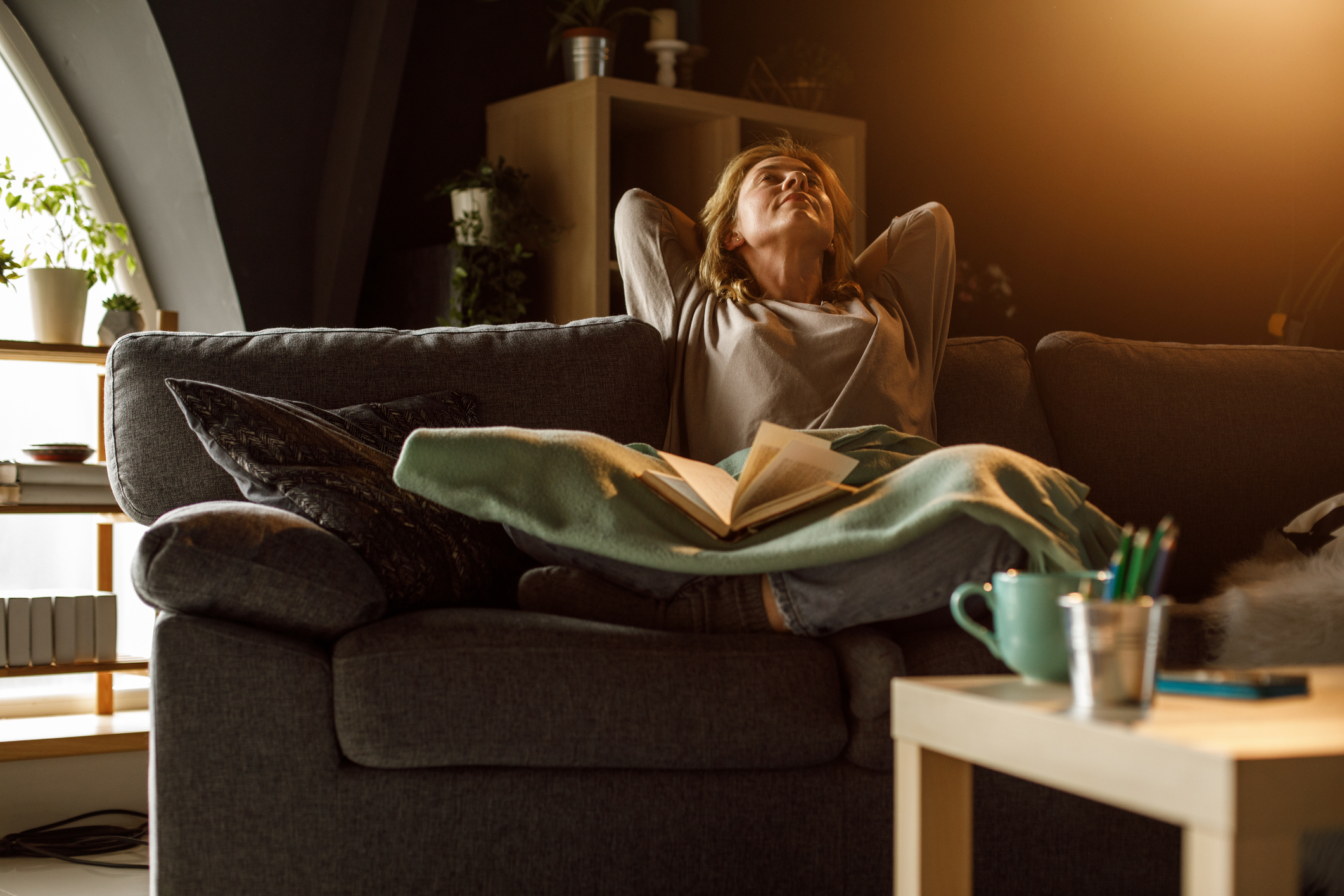 Person relaxing on a couch with hands behind their head, eyes closed, and a book on their lap. A coffee table with a mug and papers is in the foreground