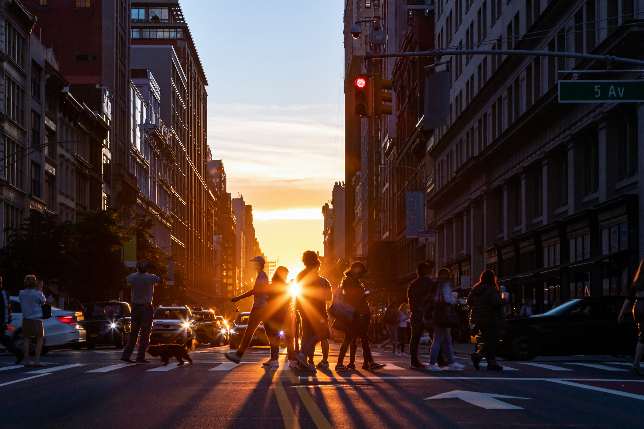 A bustling city street at sunset with pedestrians crossing as traffic waits. Buildings on either side frame the glowing sun in the background