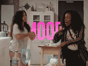 Two women dance and laugh together in a kitchen. The word "MOOD" in large letters is superimposed in the background
