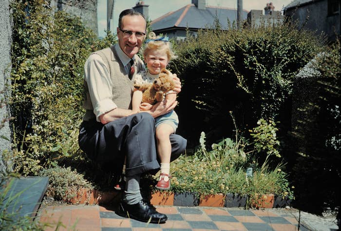 A man kneels beside a young girl holding a stuffed animal, both smiling in a garden setting with greenery and houses in the background