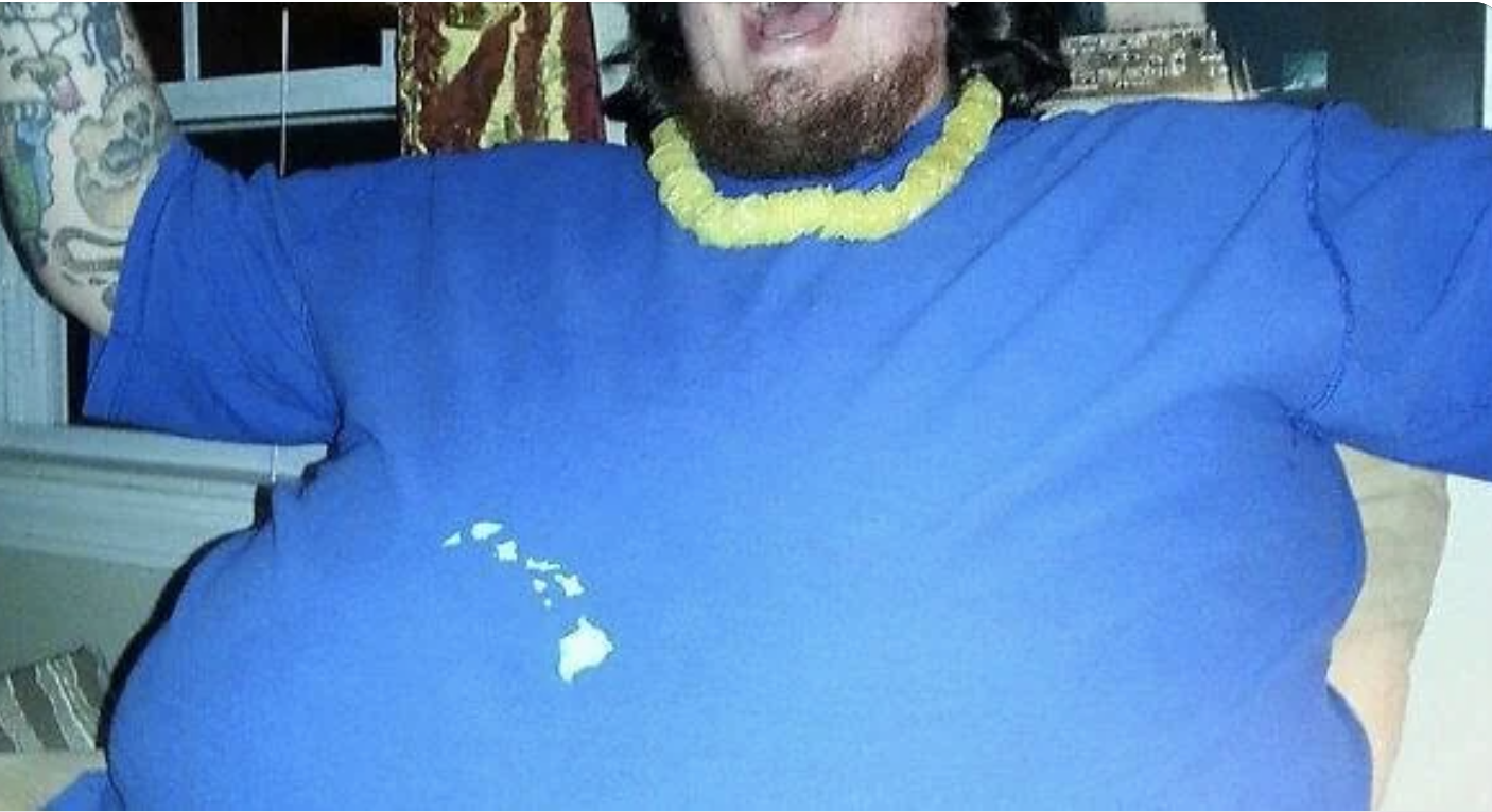 A person with tattoos and wearing a yellow lei is seen wearing a blue shirt with a stain on it; face is partially visible