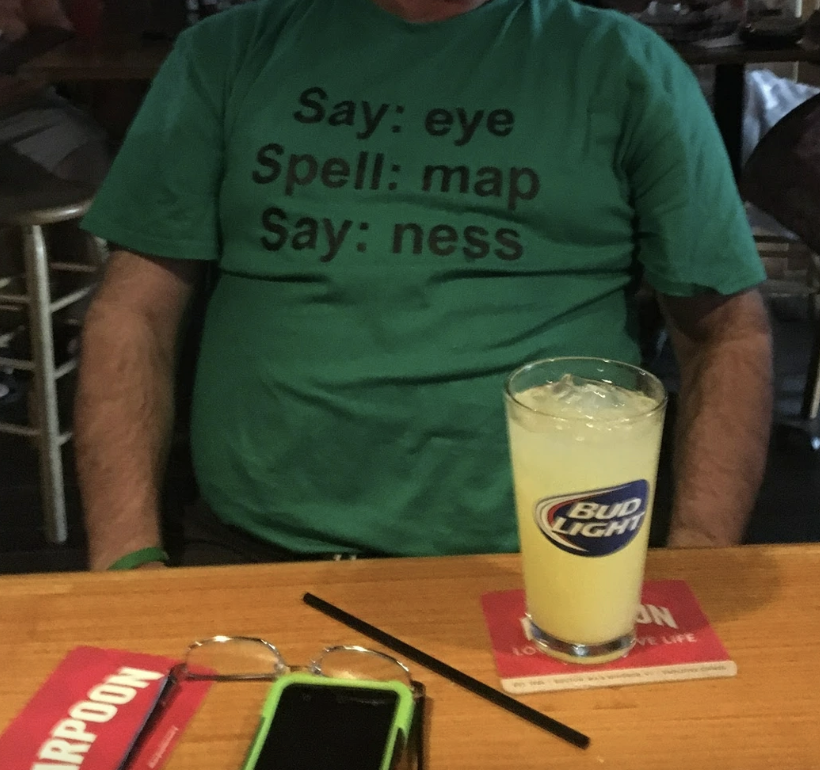 A person wearing a shirt with text: "Say: eye, Spell: map, Say: ness," seated at a table with a glass of Bud Light and a phone on the table